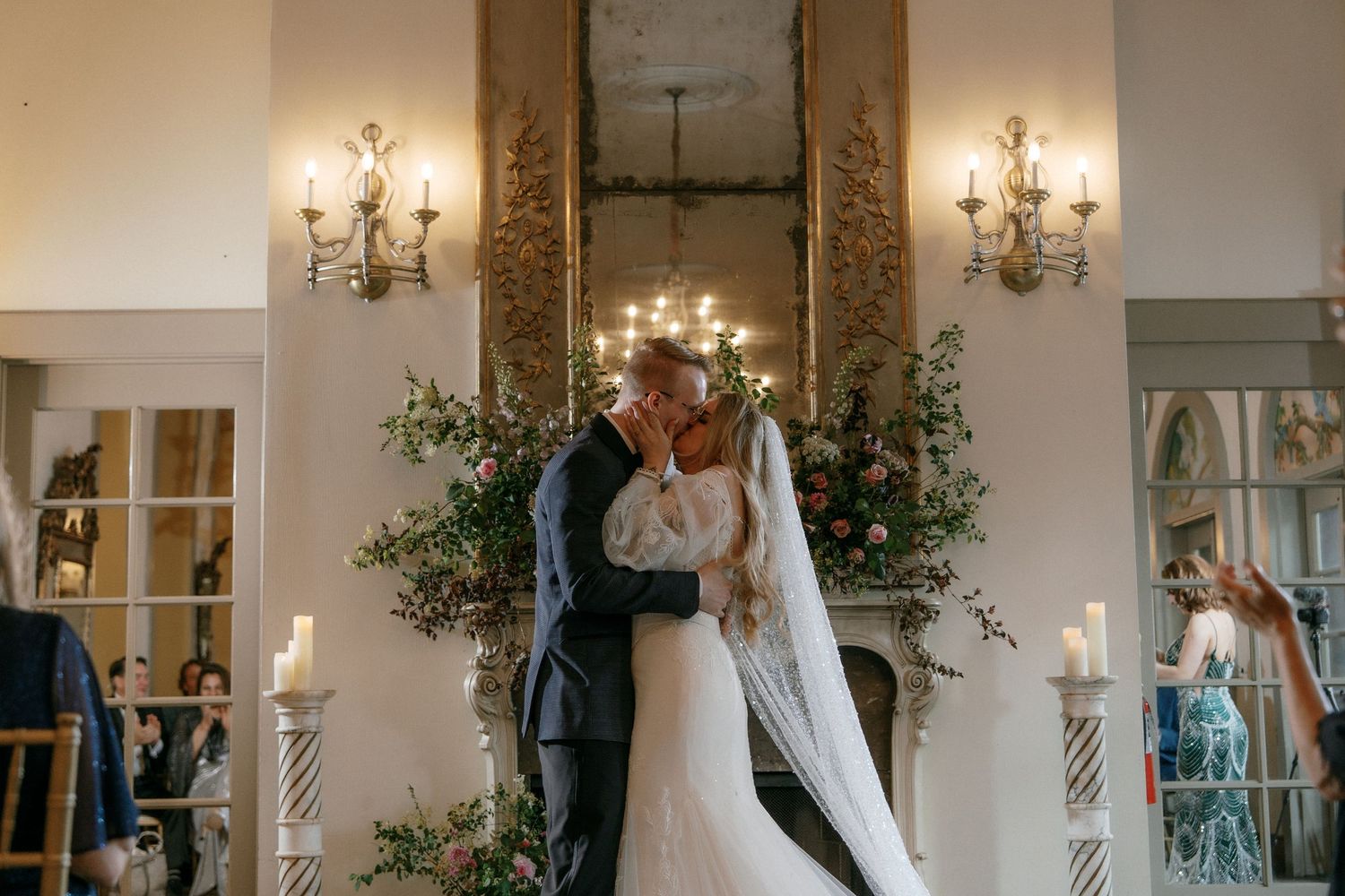 Bride and groom share first kiss before candlelit fireplace with floral arrangements at wedding.