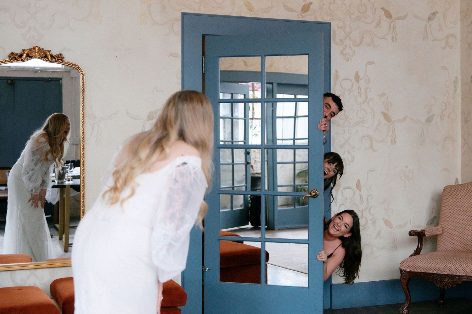 Bride in white lace gown with wedding party playfully peeking through a blue glass-paned door.