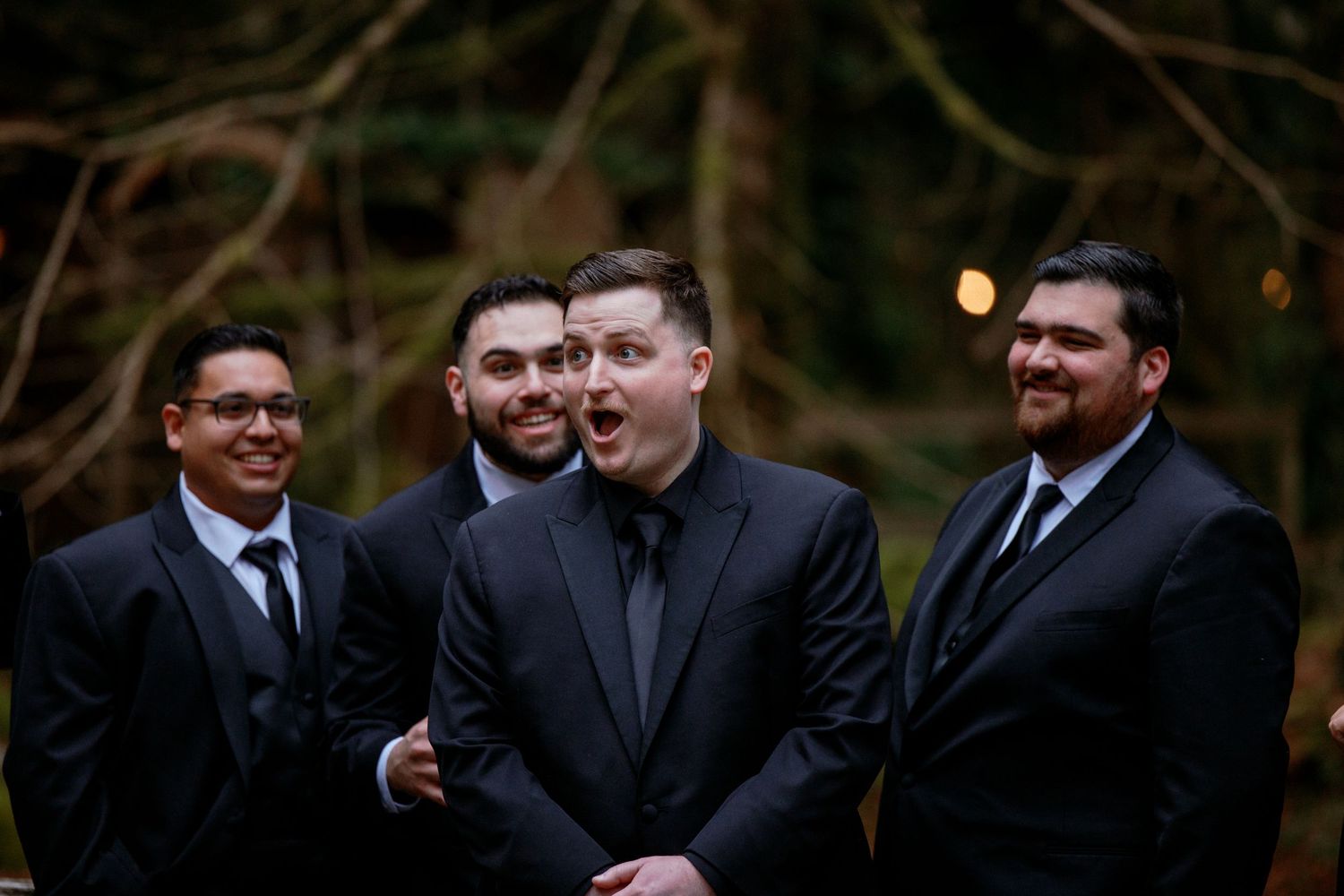Four groomsmen in black suits laughing together outdoors at a forest wedding celebration.