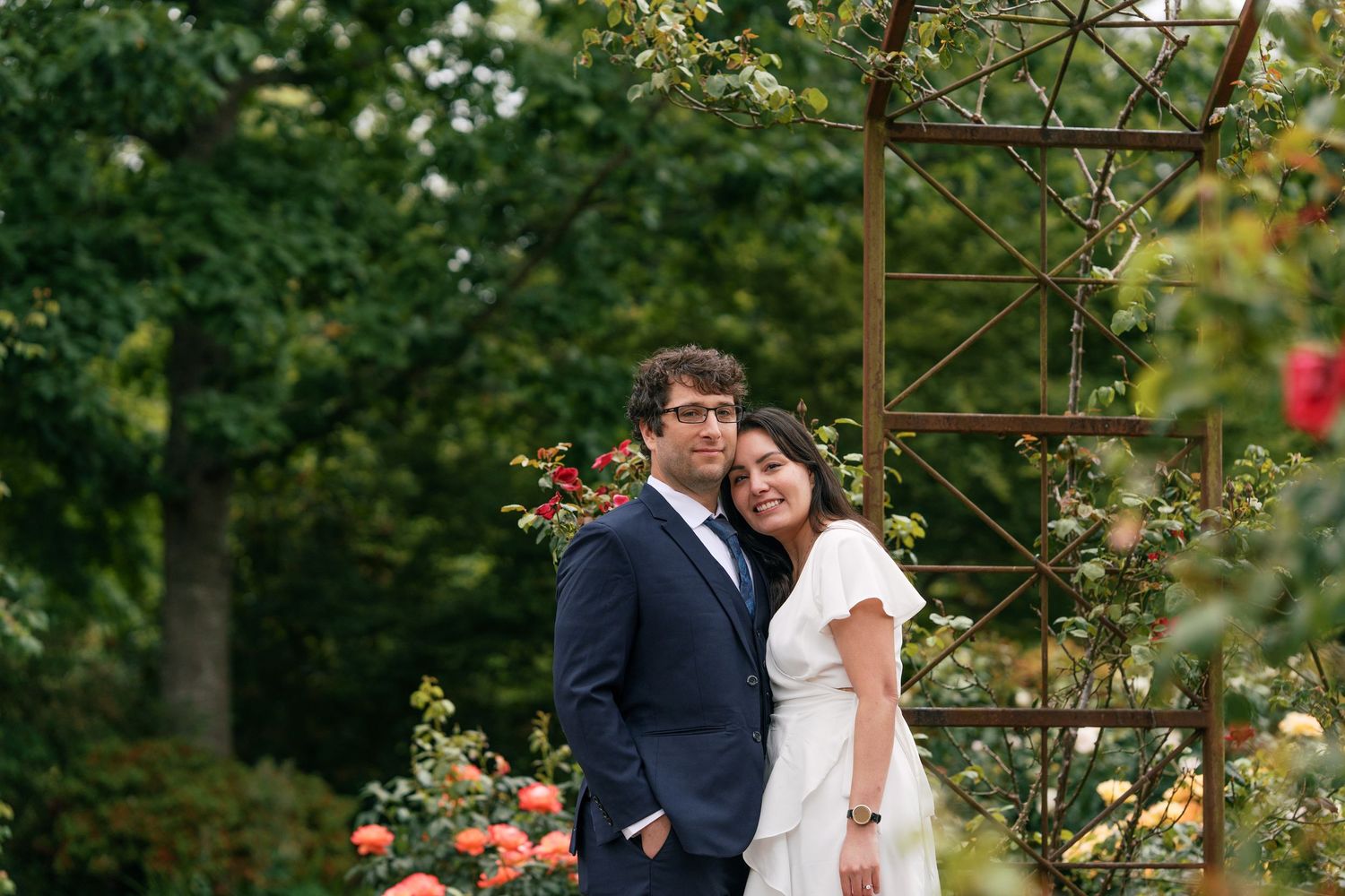 Newlywed couple poses romantically in lush rose garden beside rustic metal trellis.