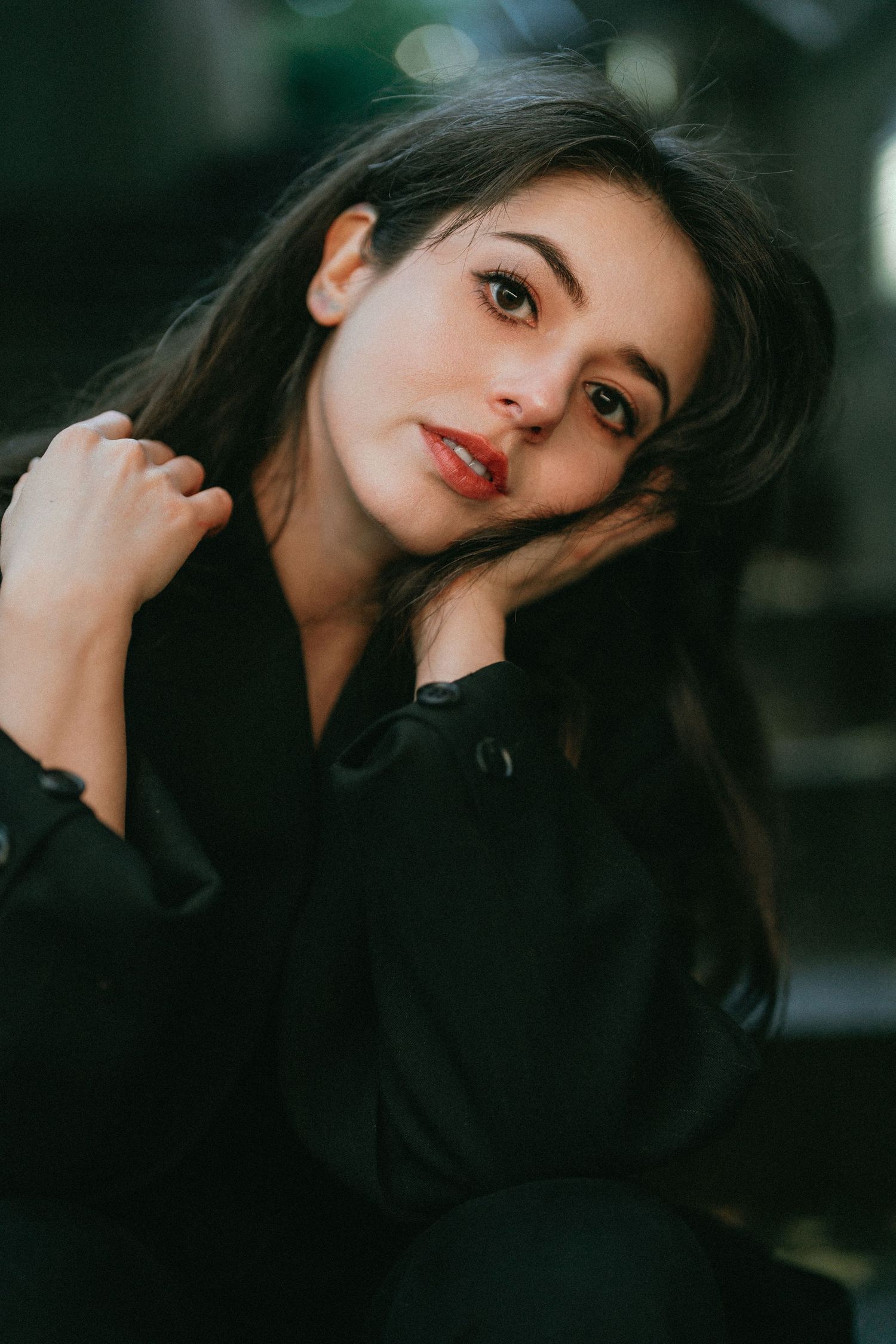Young woman with dark hair in black jacket poses thoughtfully, resting head on hand in moody portrait.