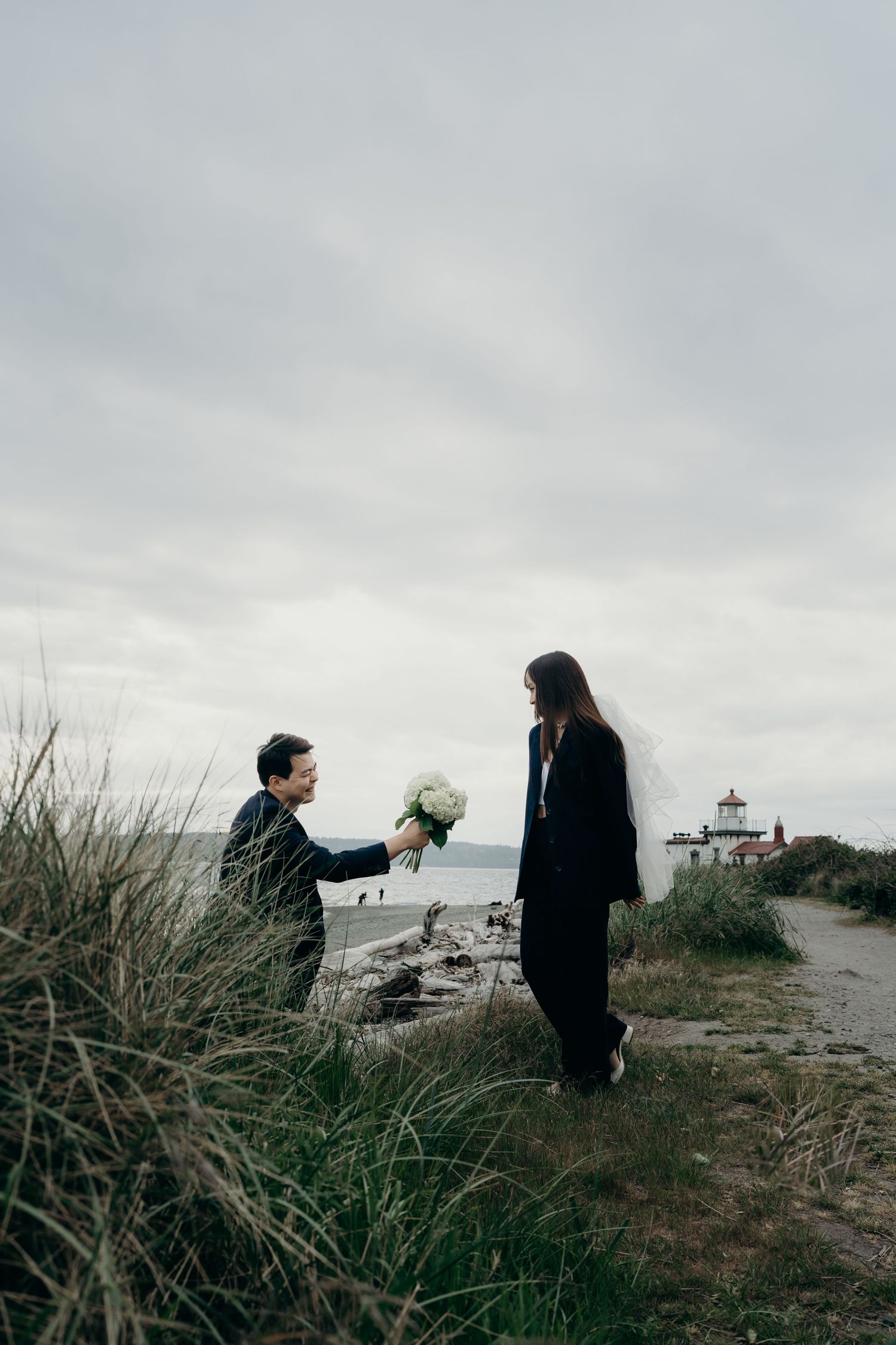 Person kneeling to present a white bouquet to a veiled bride near a coastal lighthouse.