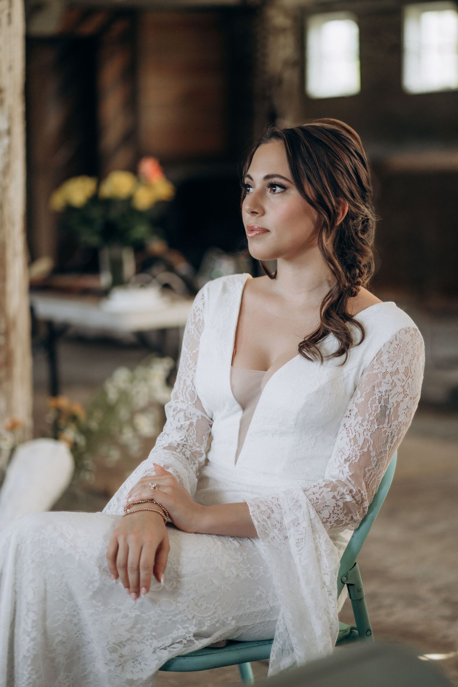 Elegant bride in white lace wedding gown seated on teal chair gazing thoughtfully in rustic barn venue.