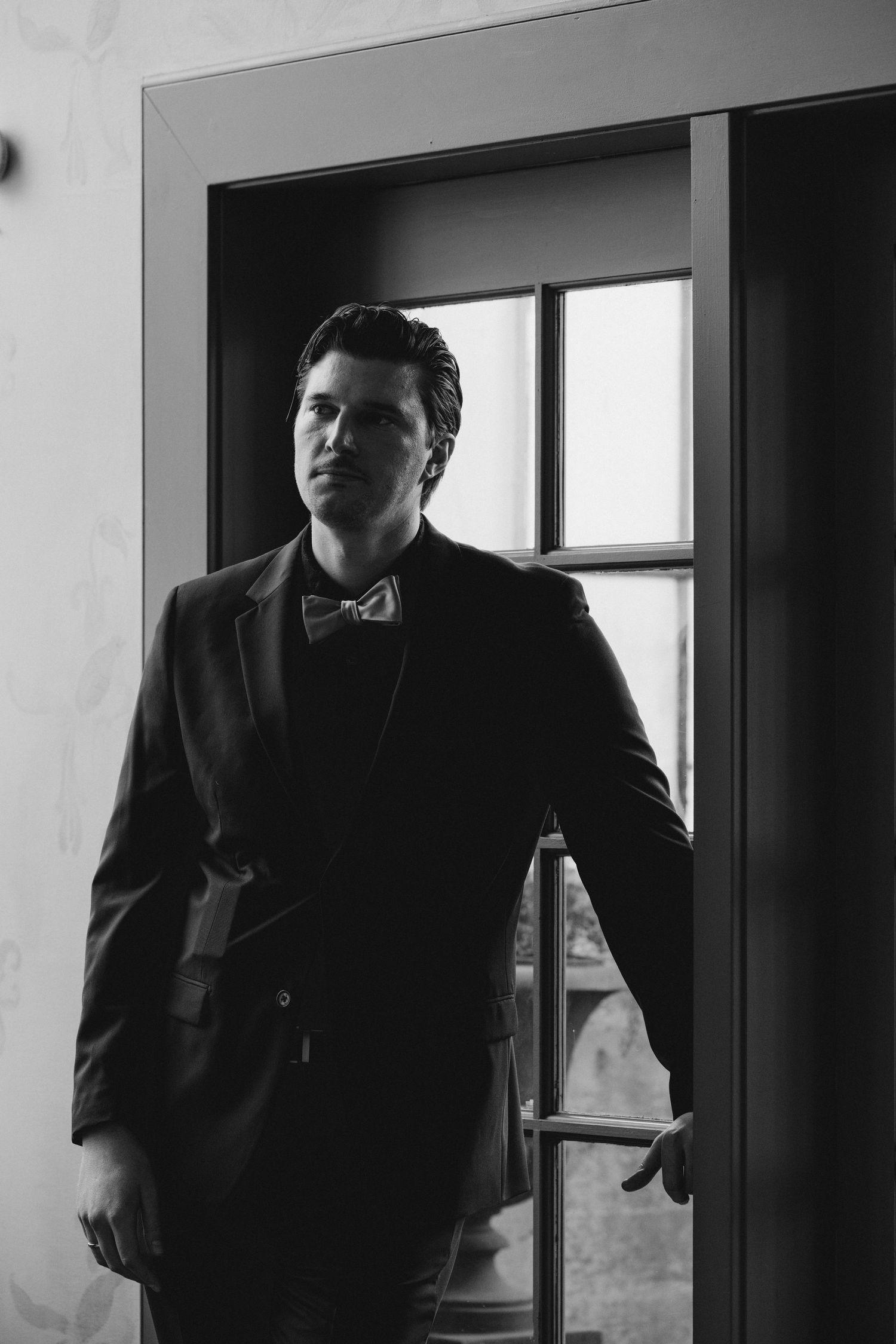 Dapper groom in black tuxedo and bow tie leaning against a doorframe in black and white portrait.