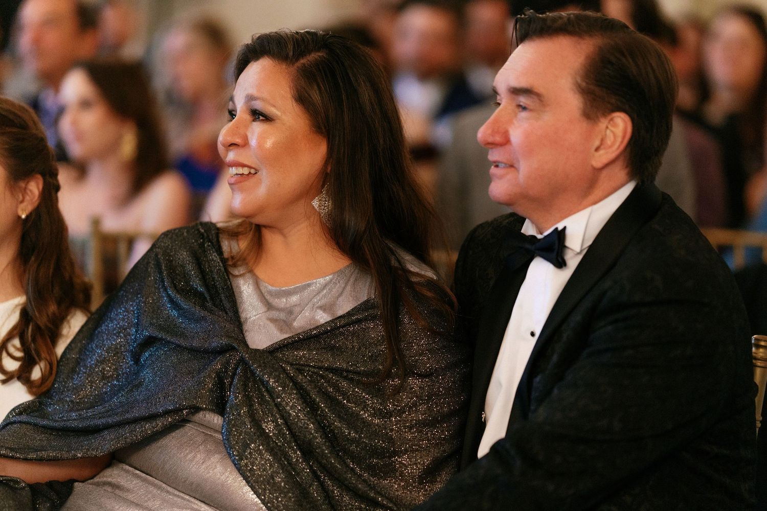 Elegant couple in formal attire seated together as guests at a wedding reception or ceremony.
