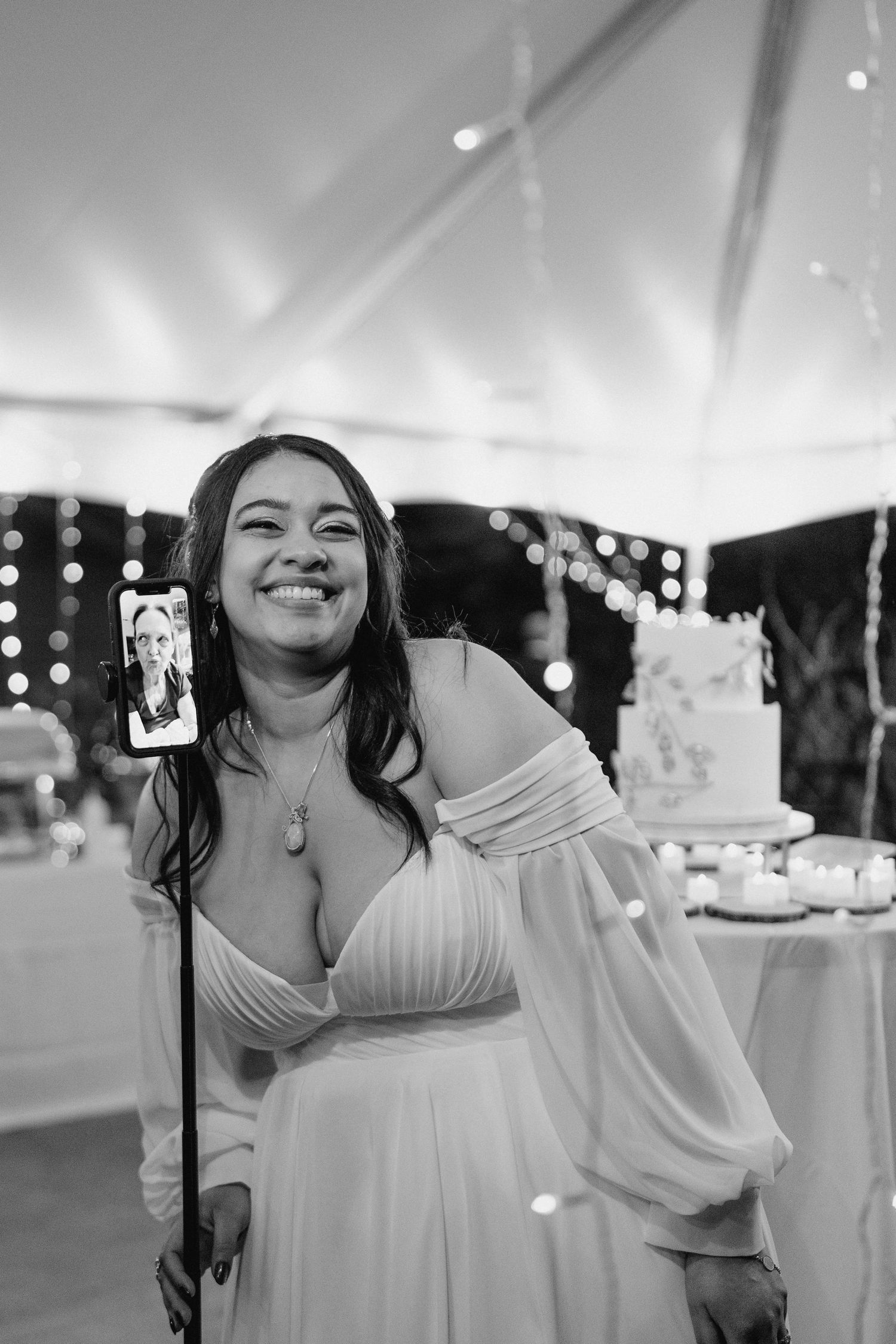 Bride in white gown smiling while video calling on phone at wedding reception with fairy lights.