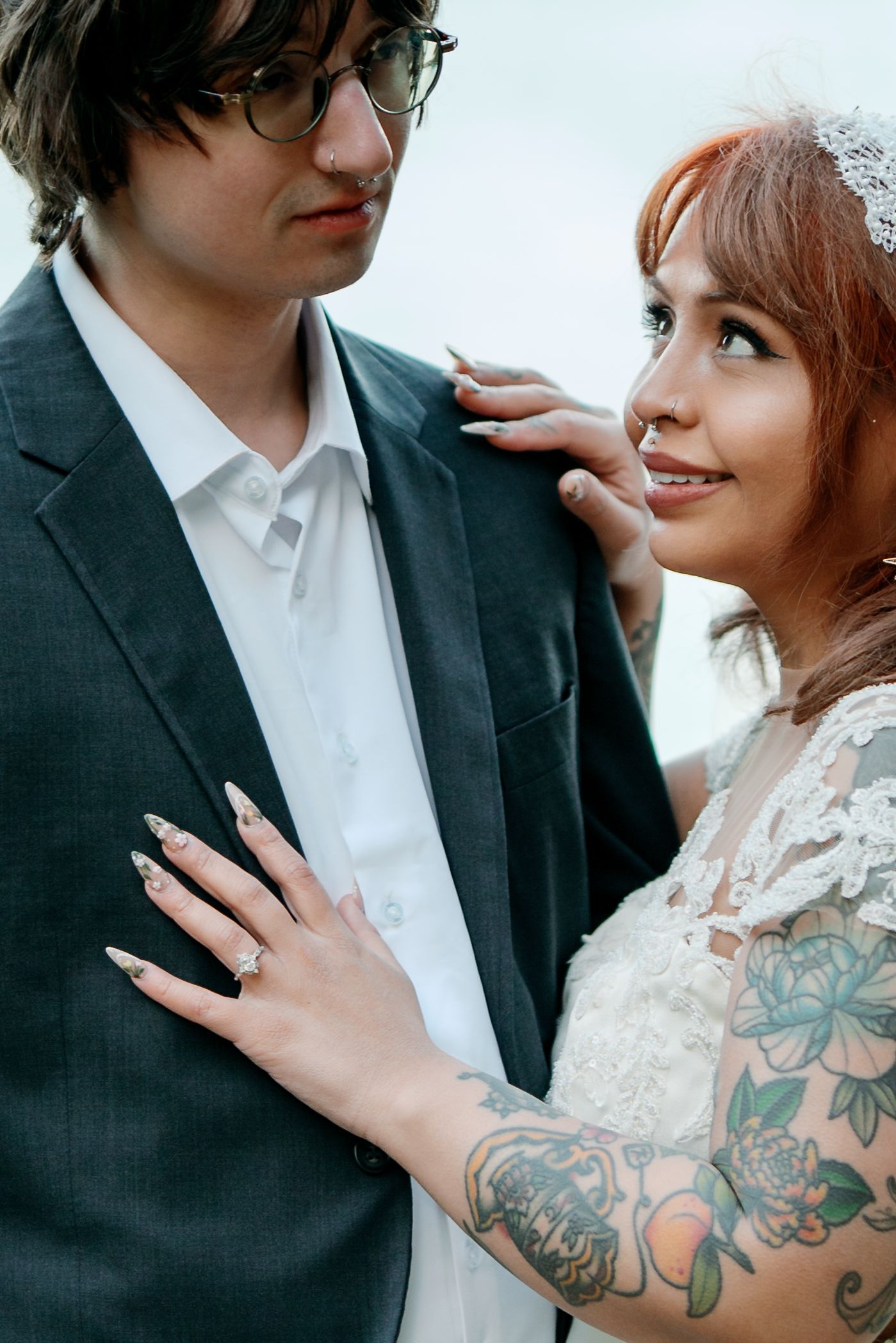 Tattooed bride in lace wedding dress gazes up at groom while showing off her diamond engagement ring.