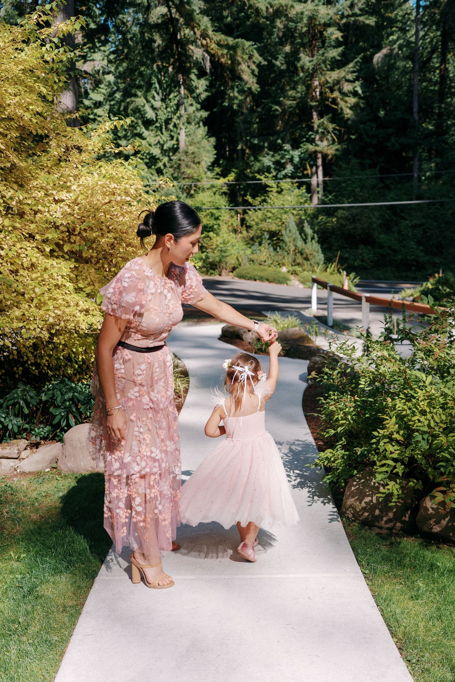 Woman in pink floral dress holding hands with young flower girl in blush tutu on garden pathway.