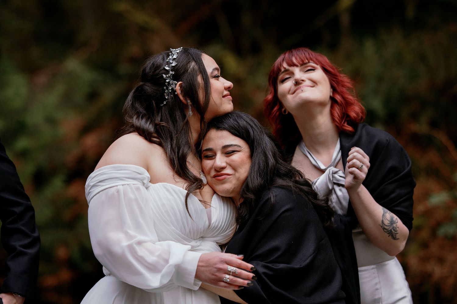 Bride in white gown sharing joyful group hug with two women at wedding.