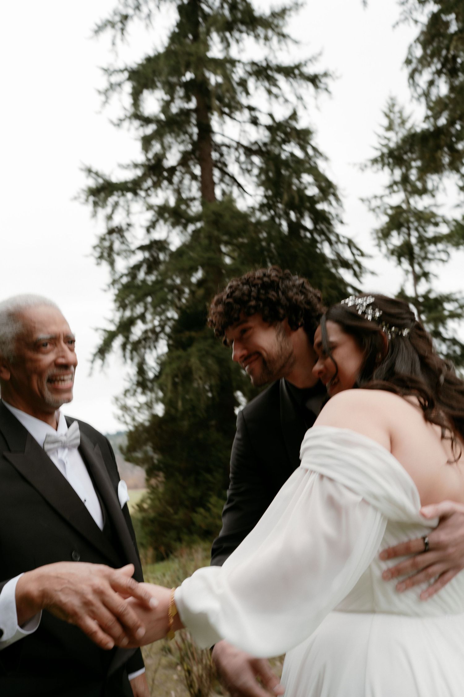 Older gentleman in tuxedo smiling as bride and groom laugh together at outdoor wedding ceremony.