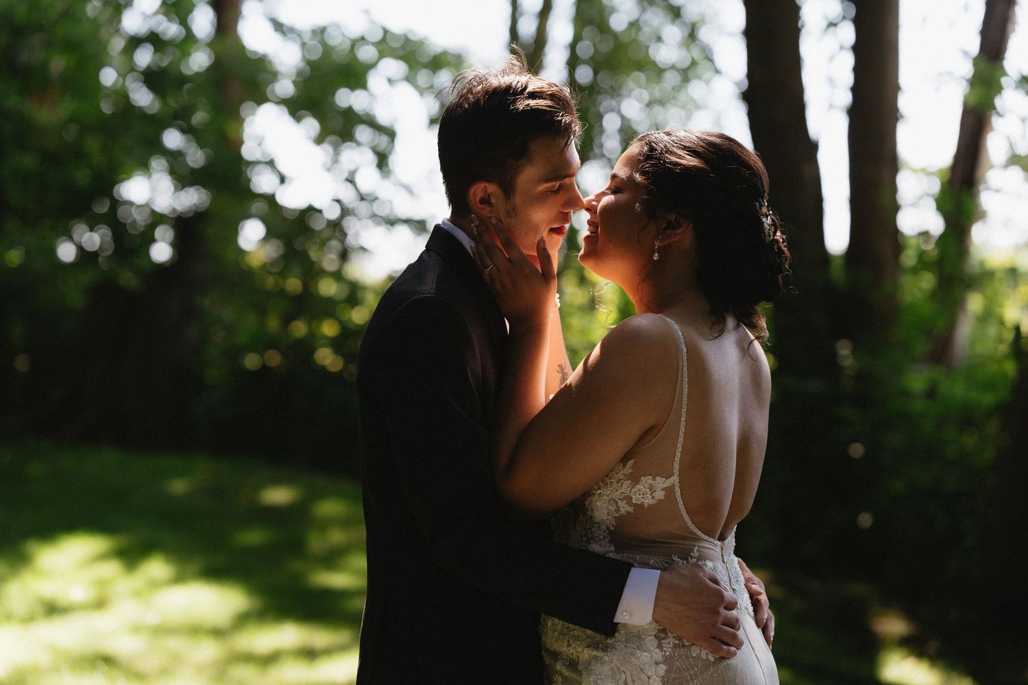 Bride and groom share an intimate moment face to face in a sunlit garden with bokeh trees.