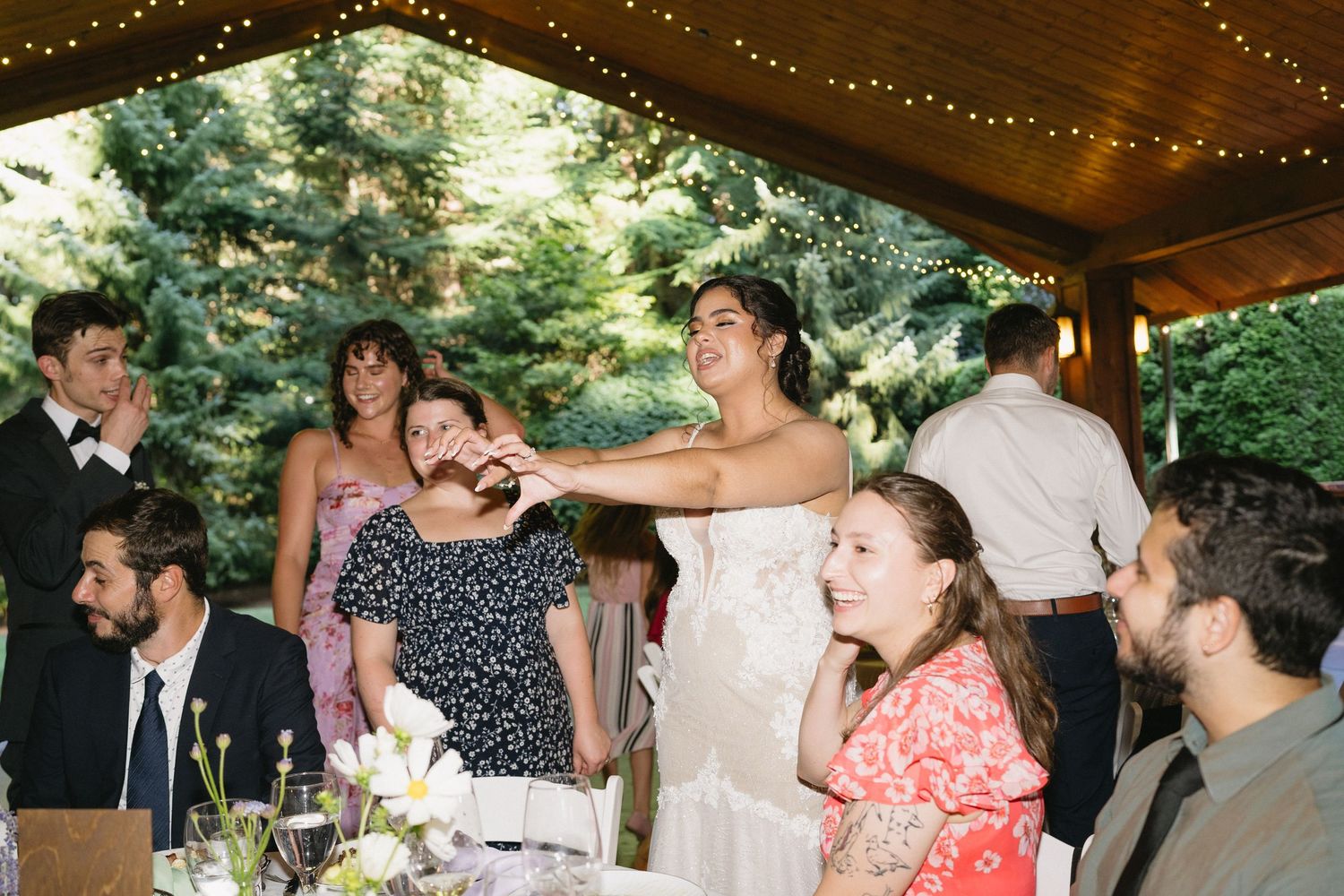 Bride in white gown interacts warmly with seated wedding guests under a rustic outdoor pavilion.