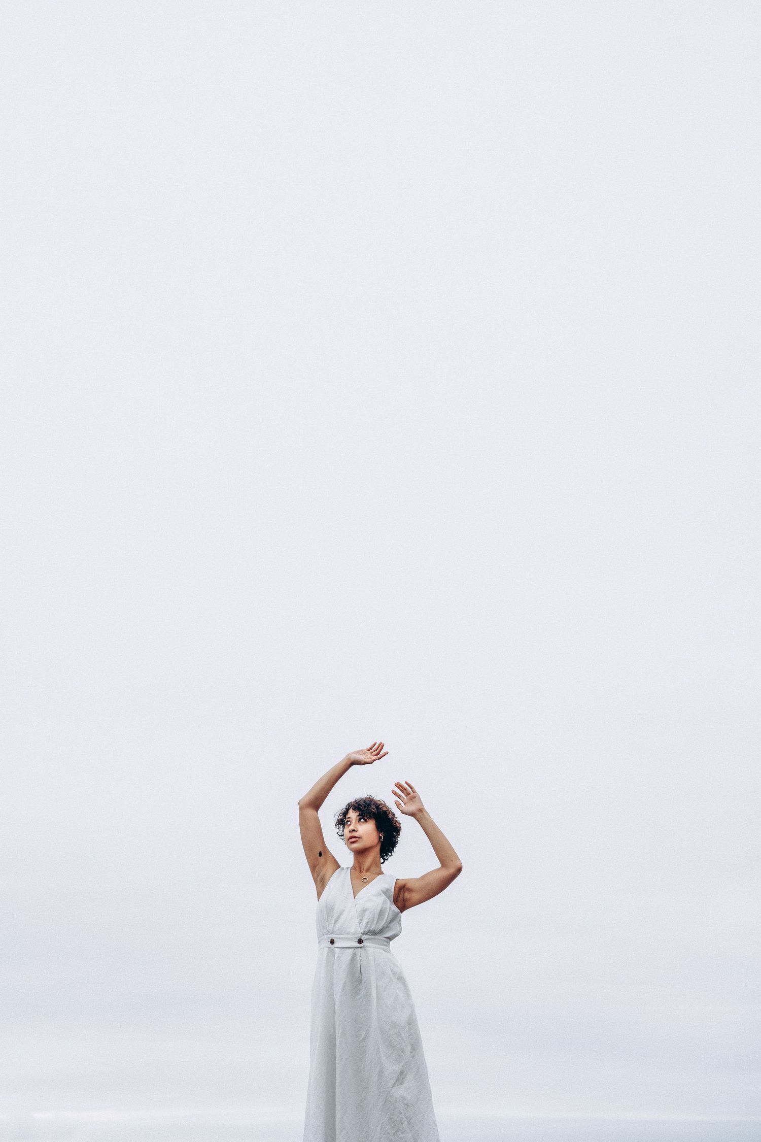 Woman in white dress with arms raised gracefully against overcast sky near the ocean.