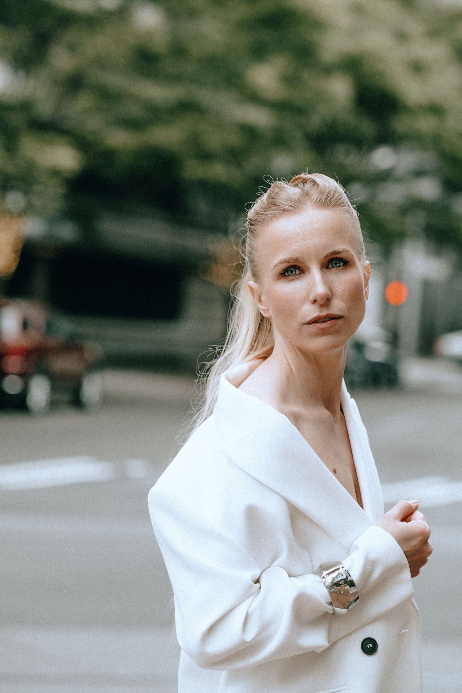 Blonde woman in white oversized blazer and silver watch poses on urban street with blurred background.