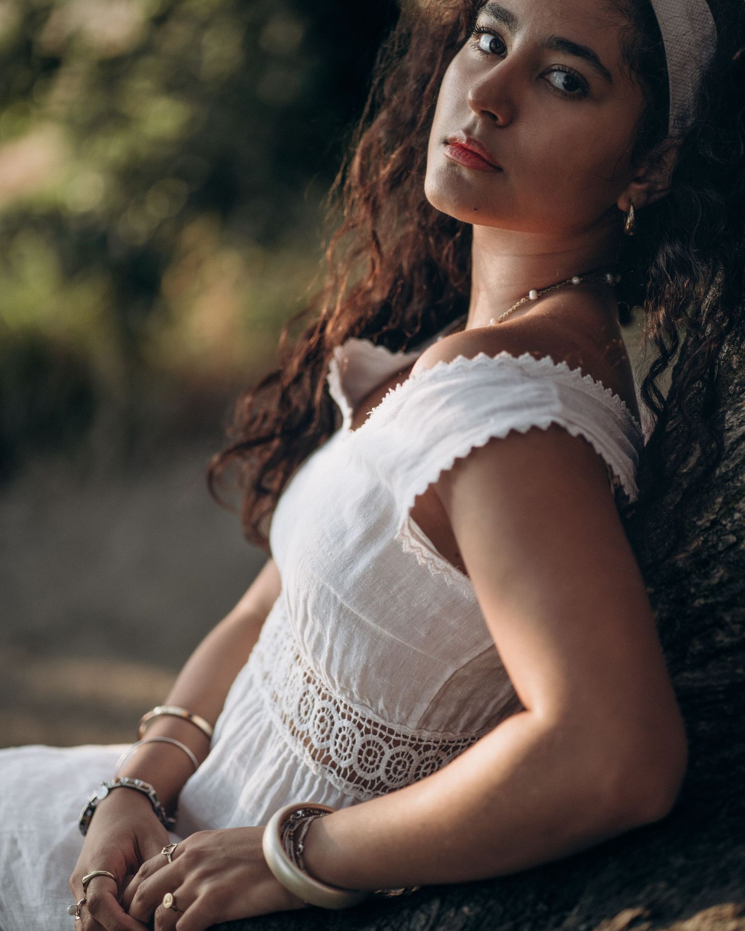 Young woman in white off-shoulder lace dress with curly hair posing outdoors in sunlight.