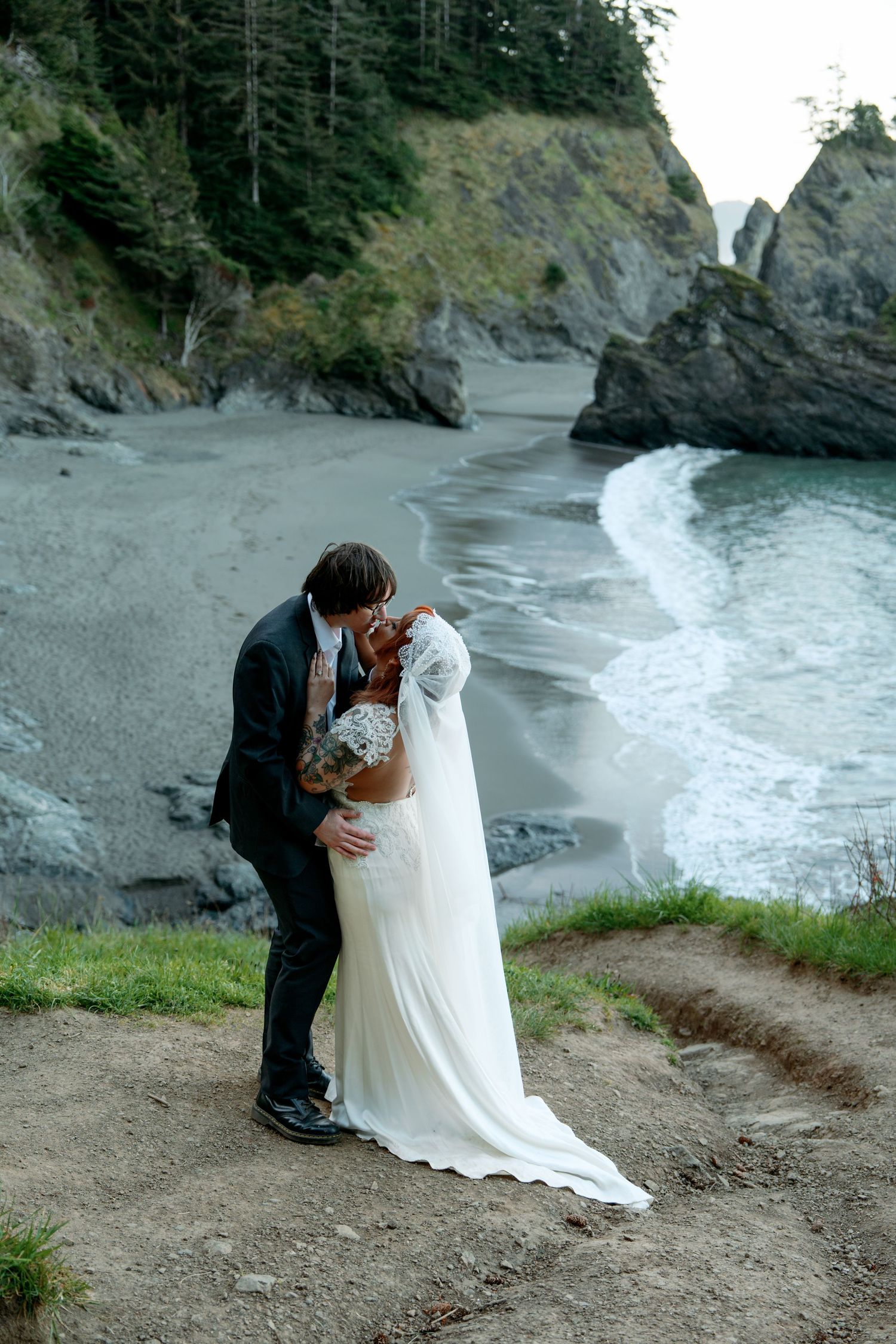 Bride and groom sharing a passionate kiss on coastal cliffs with sandy beach and waves below.
