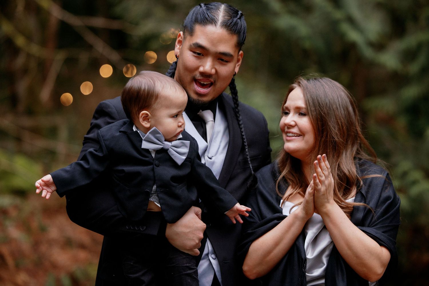 Young family smiling outdoors at wedding, man holding baby in tuxedo with woman in silver dress.