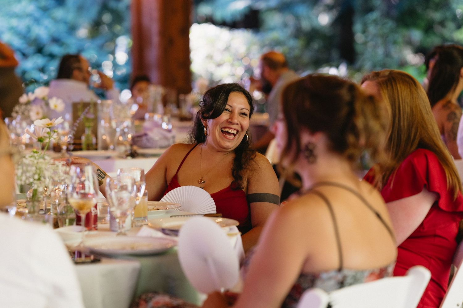 Wedding guest in red dress laughs joyfully at elegant outdoor reception dinner table.