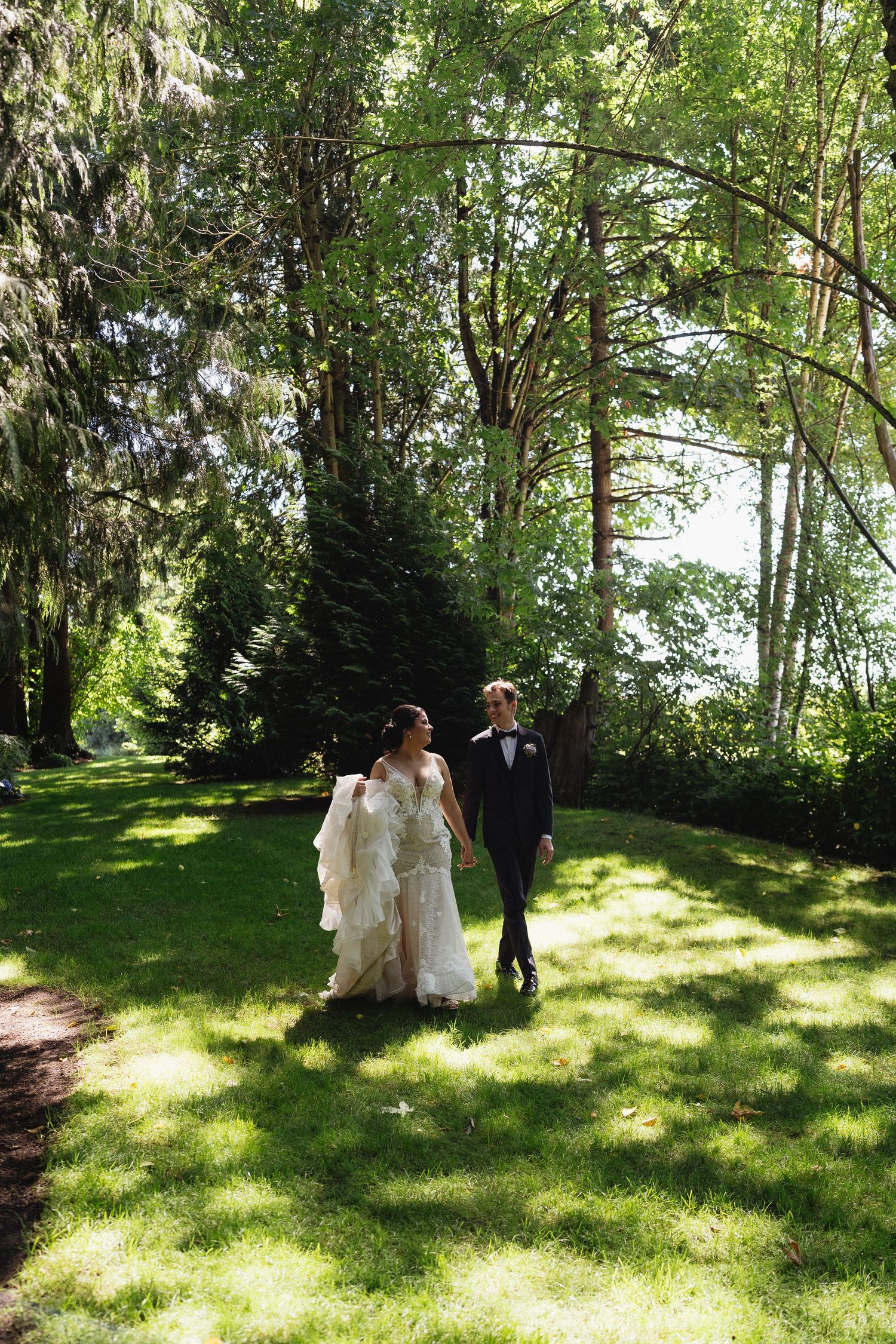 Bride and groom walk hand in hand through a sunlit garden path lined with tall green trees.