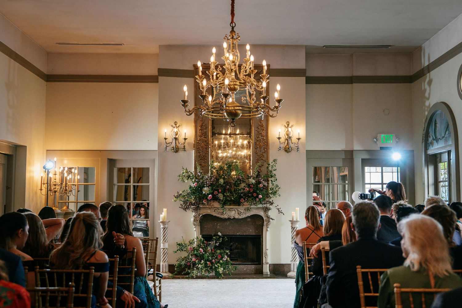 Elegant indoor wedding ceremony with guests seated facing a floral fireplace altar under a gold chandelier.