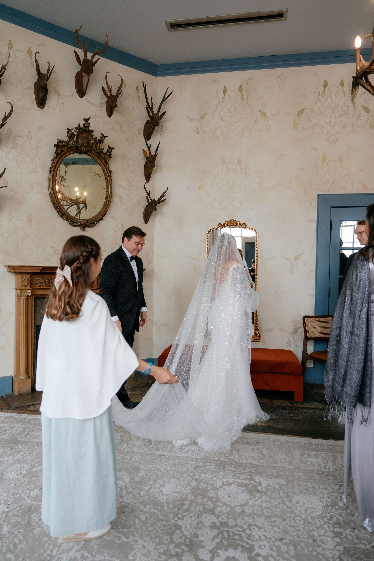 Bride in a long veil being assisted by a bridesmaid in an elegant decorated room.