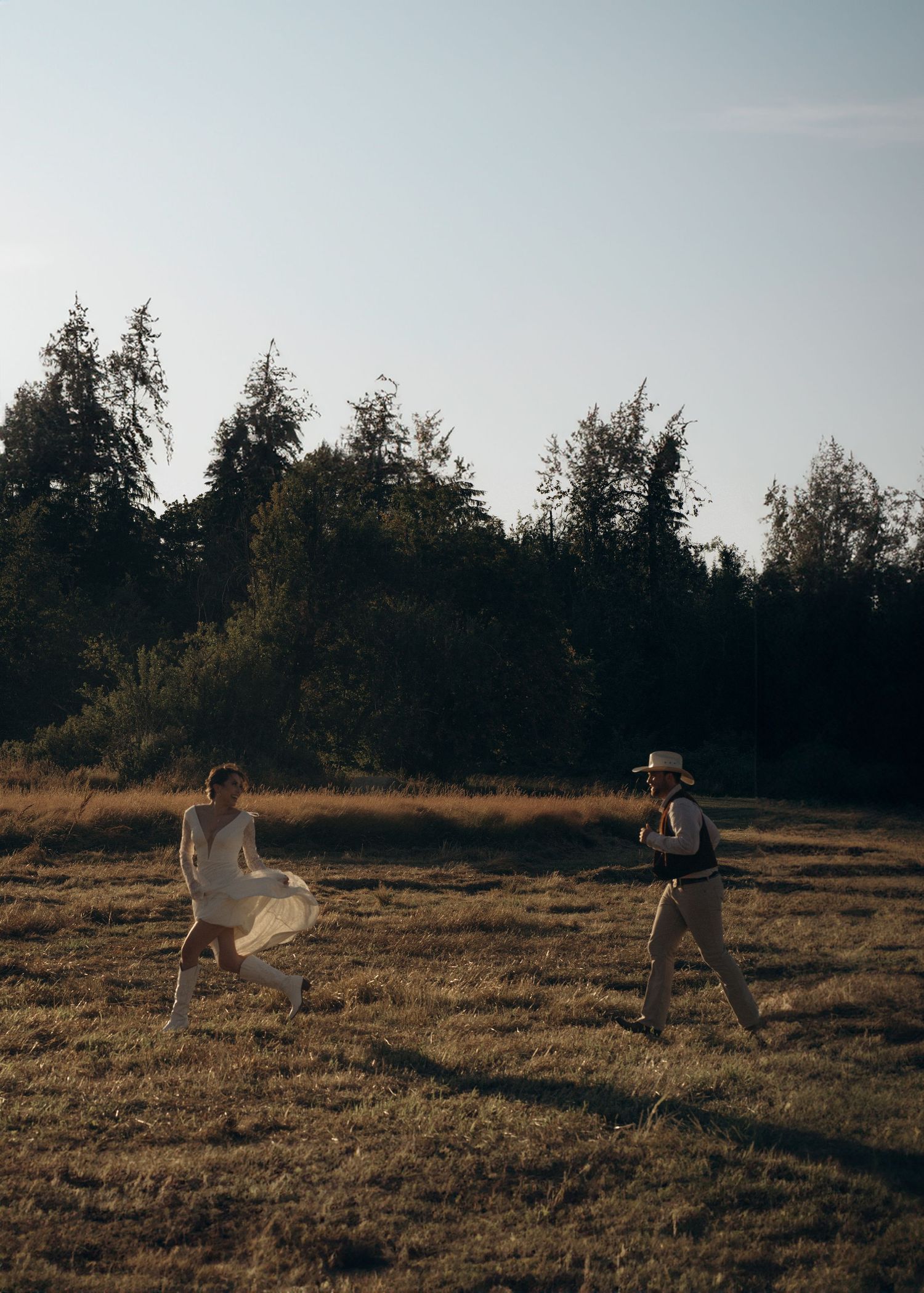 Western-styled bride and groom chasing each other across rustic field at golden hour.