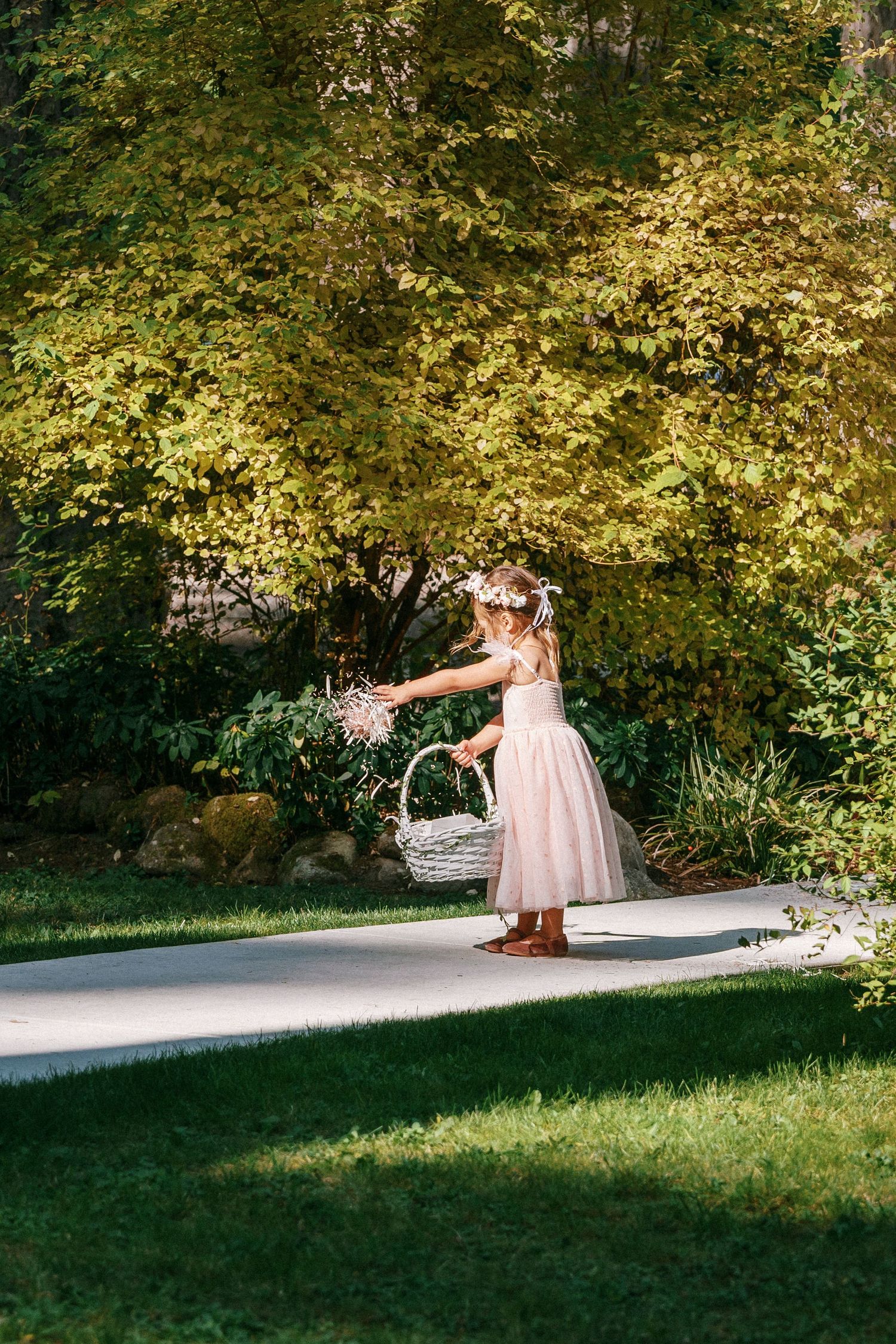 Young flower girl in pink dress and floral crown tosses petals from white basket on garden pathway.