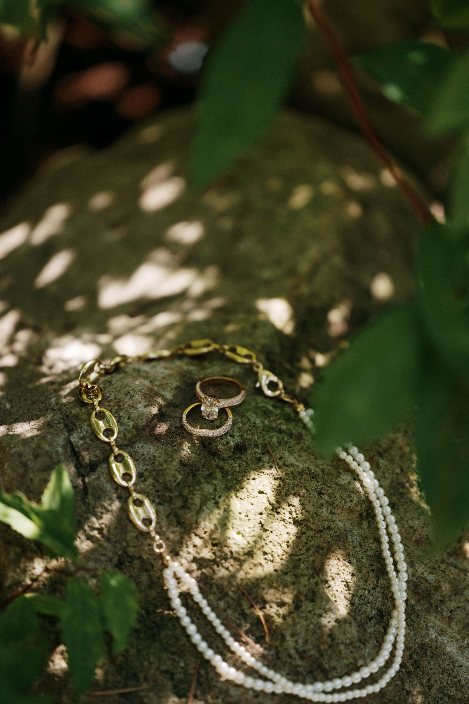 Wedding rings and pearl necklace with gold chain bracelet arranged on a moss-covered stone outdoors.