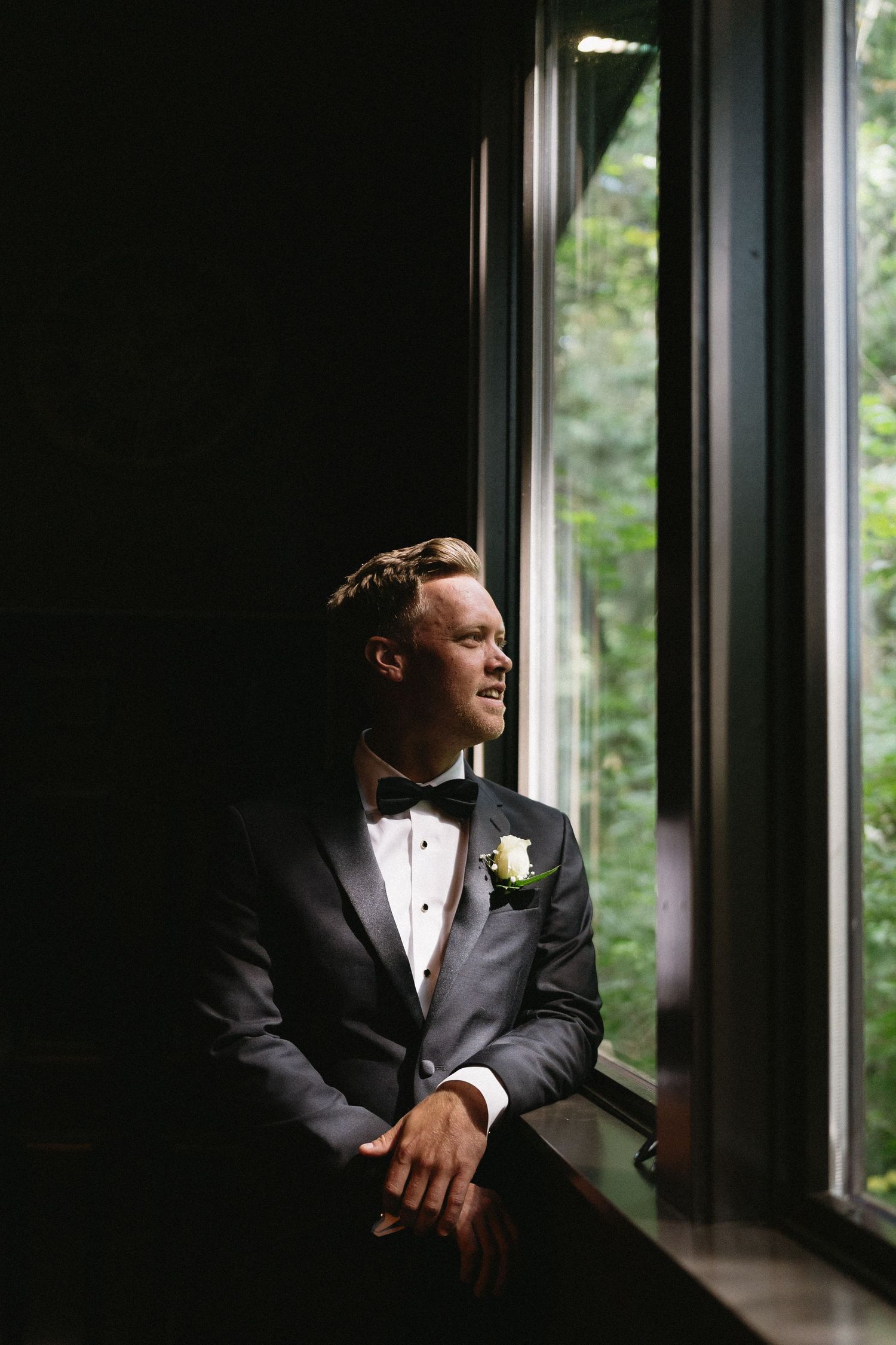 Groom in black tuxedo with boutonniere gazes thoughtfully out a large window in a softly lit room.