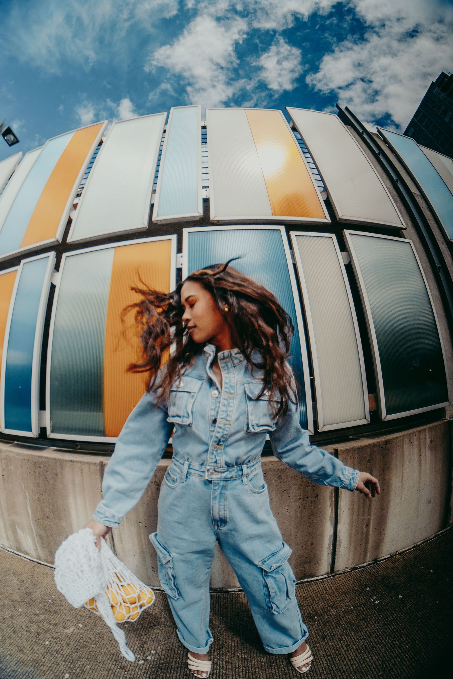 Woman in denim jumpsuit tosses her hair in front of a colorful striped building facade under blue sky.