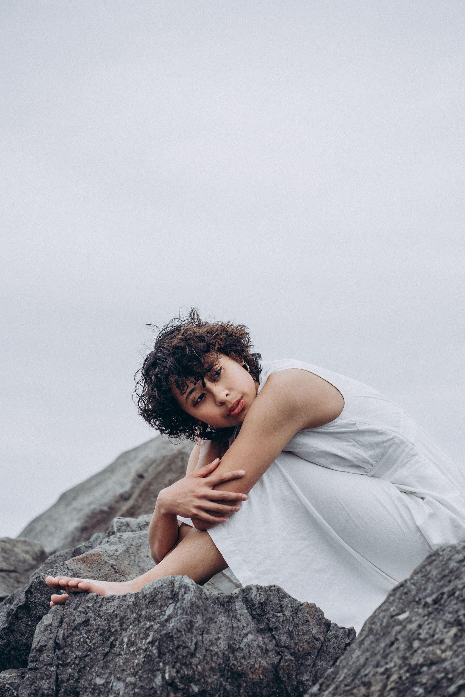 Woman in white dress sits on rocky shore hugging her knees under overcast sky.