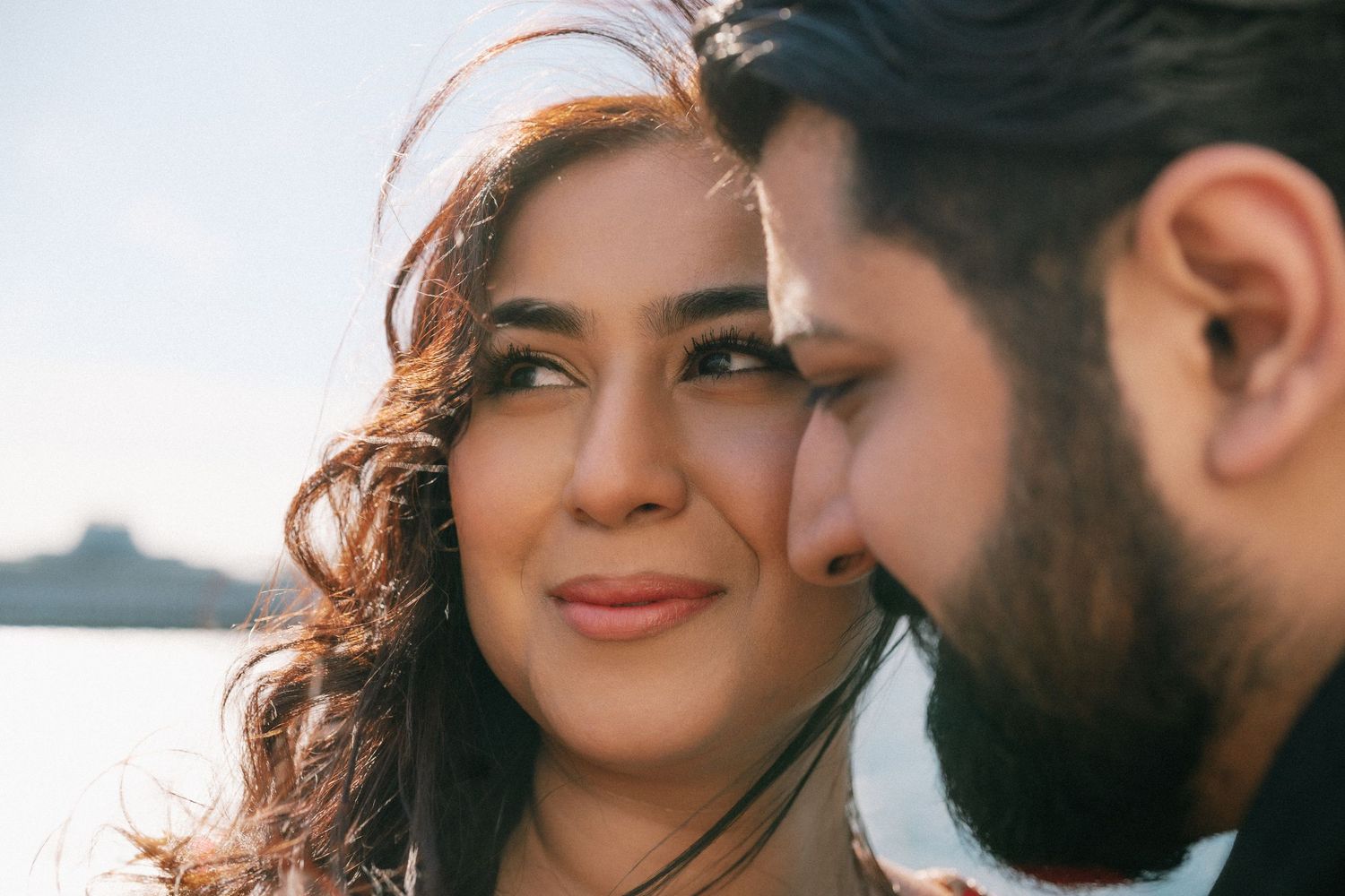 Close-up of a smiling woman and bearded man sharing an intimate moment outdoors near water.