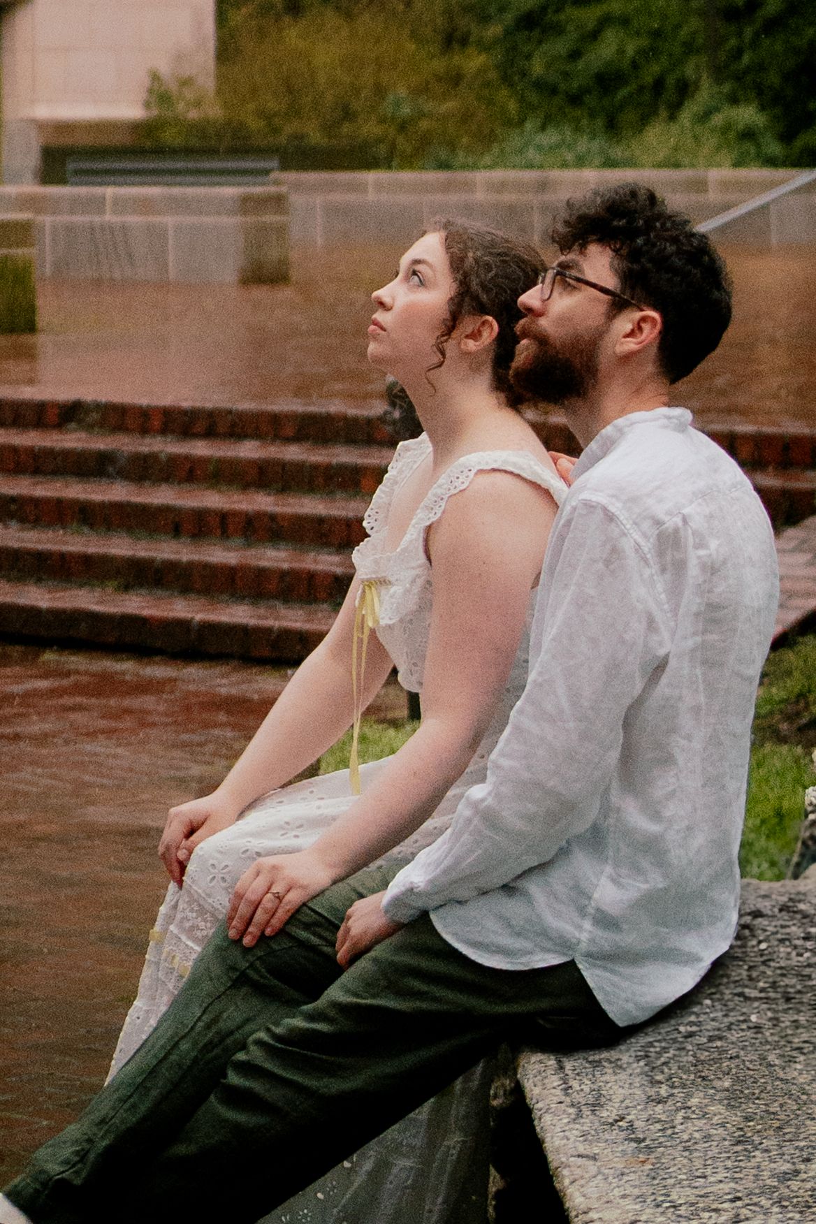 Couple sitting closely together on a stone ledge in the rain near brick steps outdoors.