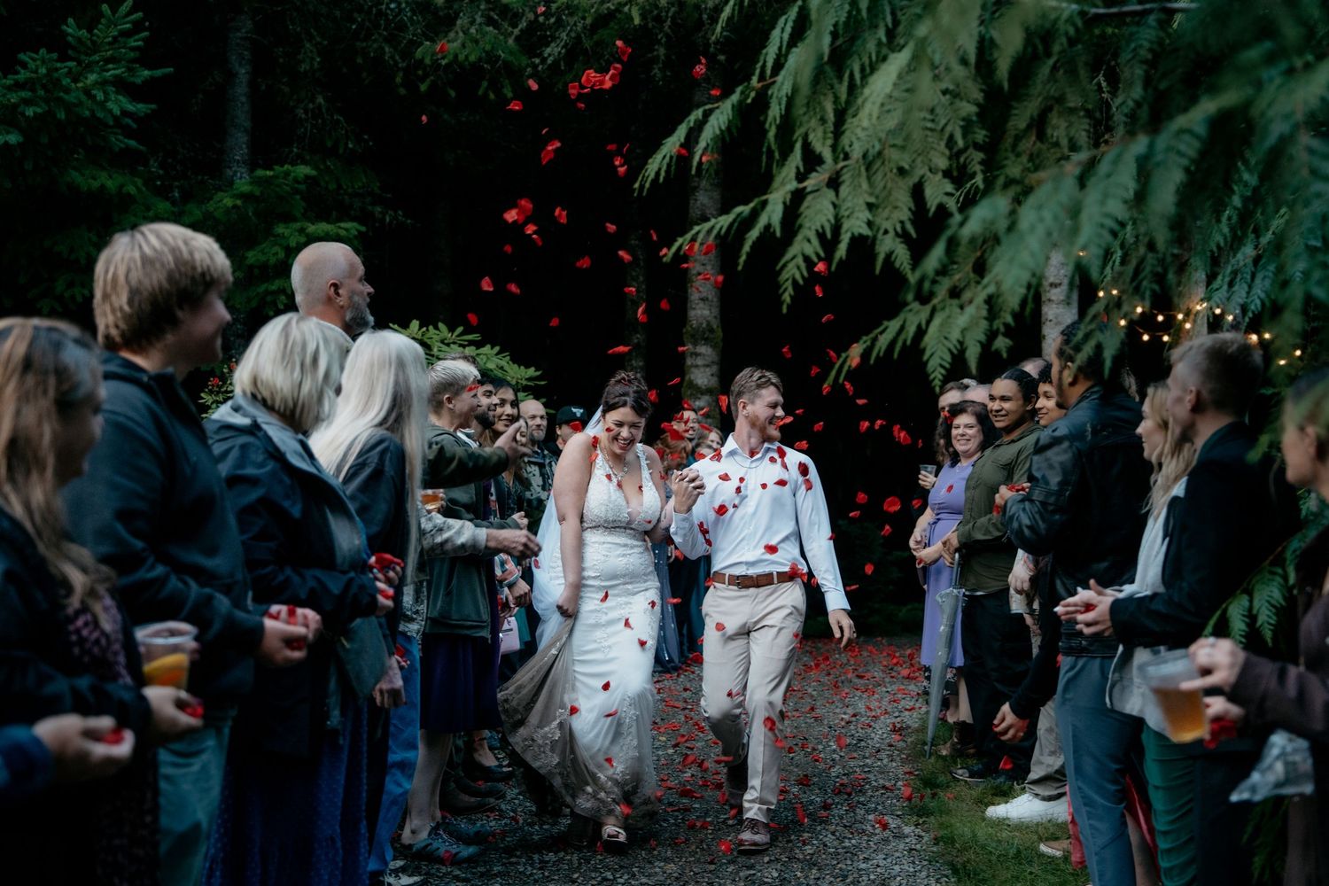 Newlywed couple walking through cheering guests tossing red flower petals in a forest wedding celebration.