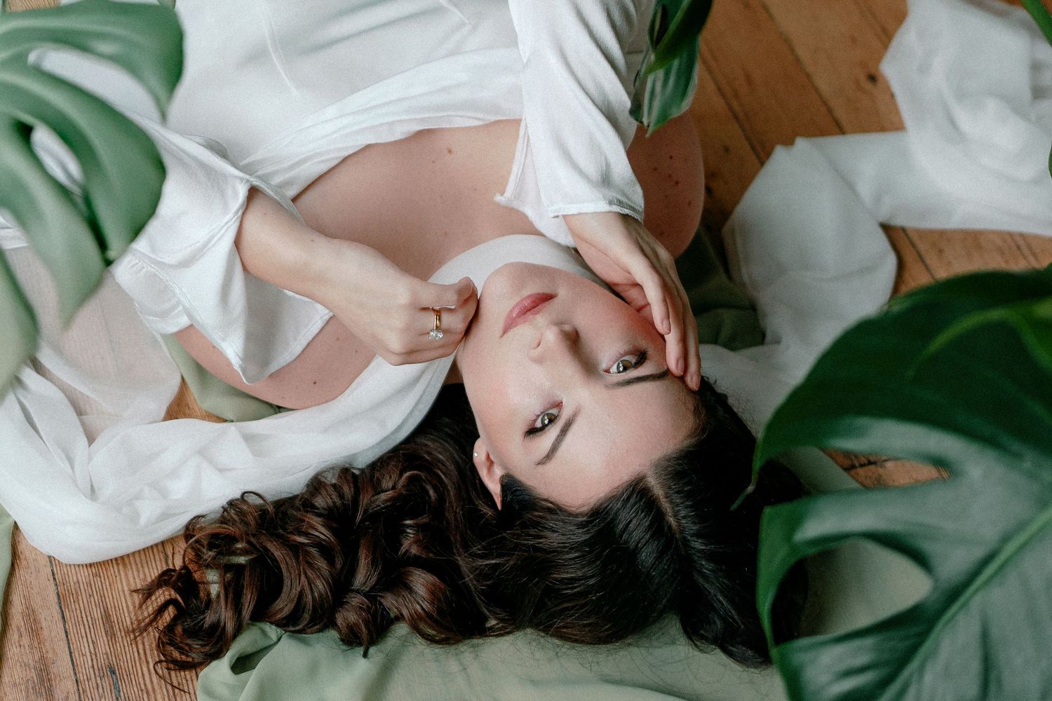 Pregnant woman in white robe lying on floor surrounded by tropical monstera leaves in a serene studio setting.