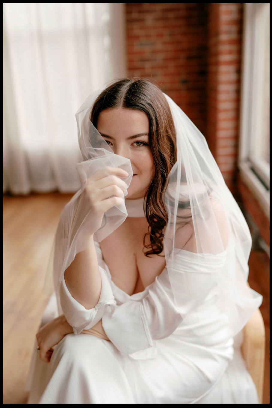 A bride in a white gown and veil holds the tulle to her lips, seated near a brick wall in soft natural light.