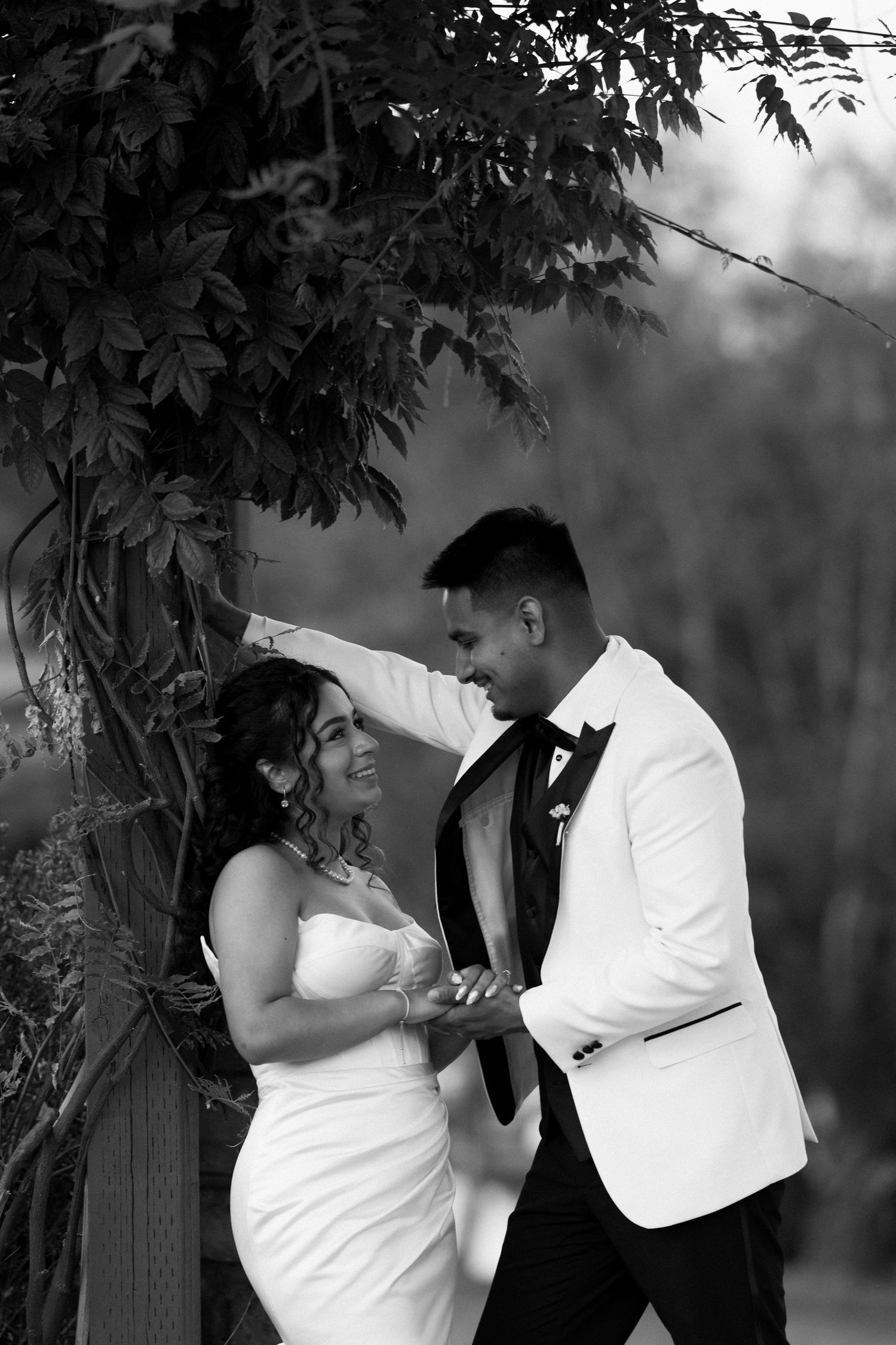 Black and white portrait of bride and groom laughing together under a leafy vine-covered wedding arbor.