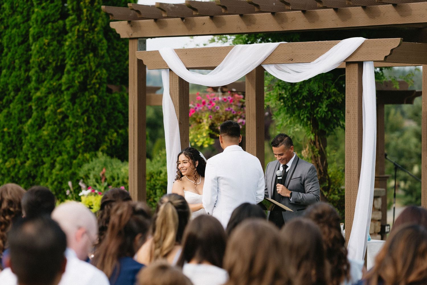 Wedding guests watch the bride and groom exchange vows under a draped pergola at an outdoor ceremony.