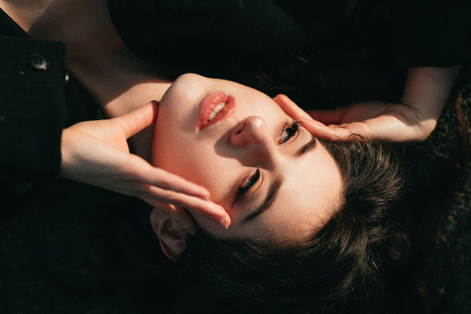 Artistic close-up of young woman lying back with hands framing face in dramatic sunlight.