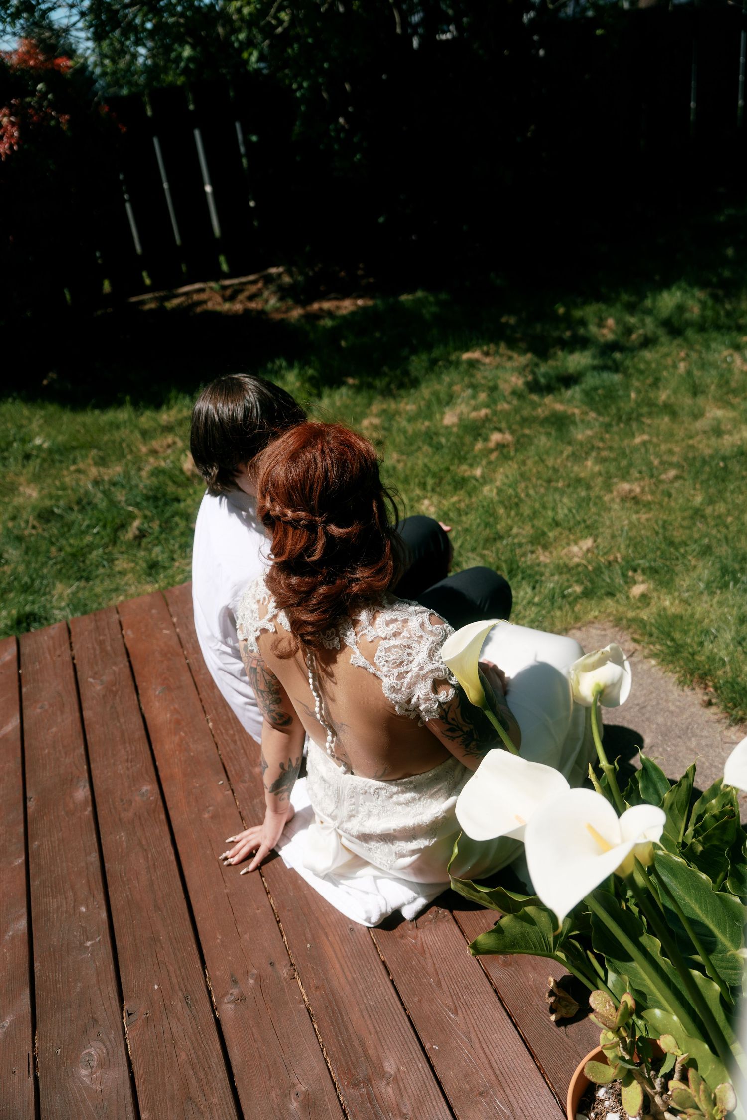 Overhead view of bride in lace dress and groom sitting on wooden deck with calla lilies.