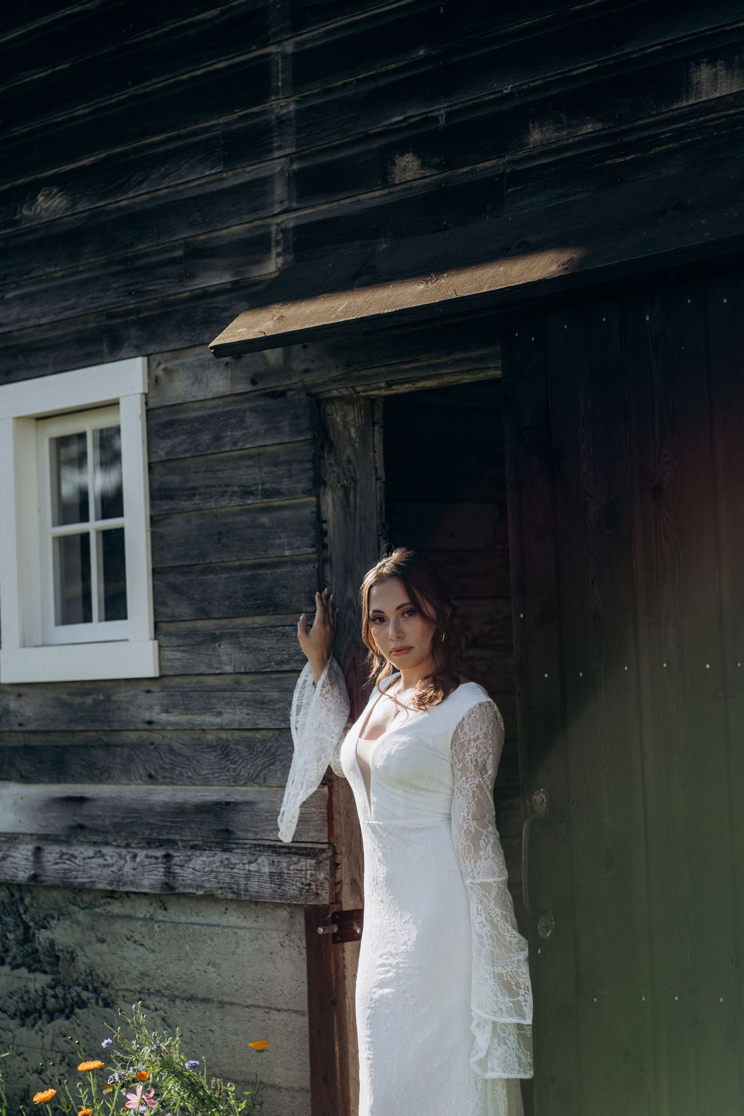 Bride in elegant white lace gown posing by weathered wooden barn door in sunlight.