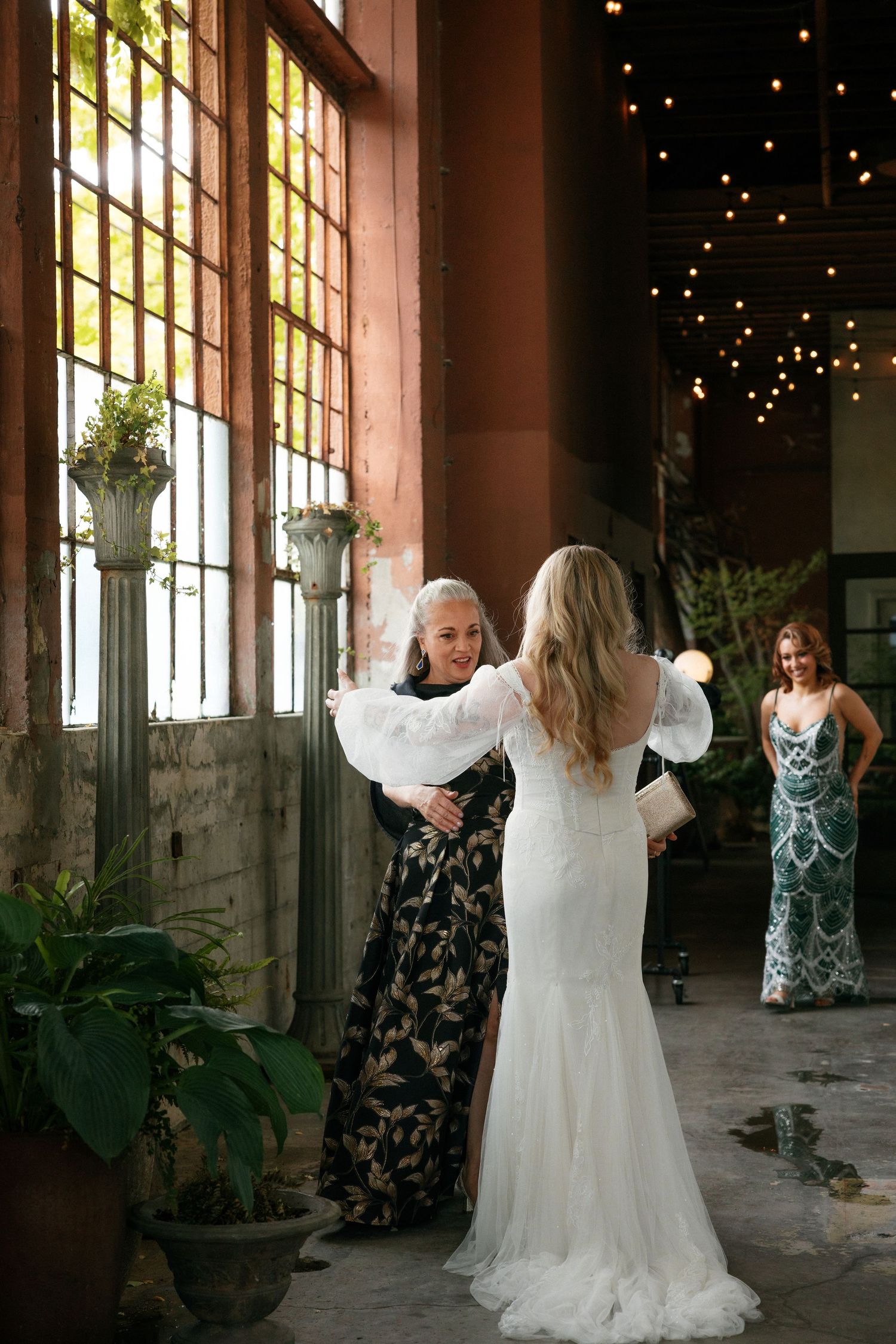 Bride greets wedding guests in an elegant industrial venue adorned with string lights and plants.
