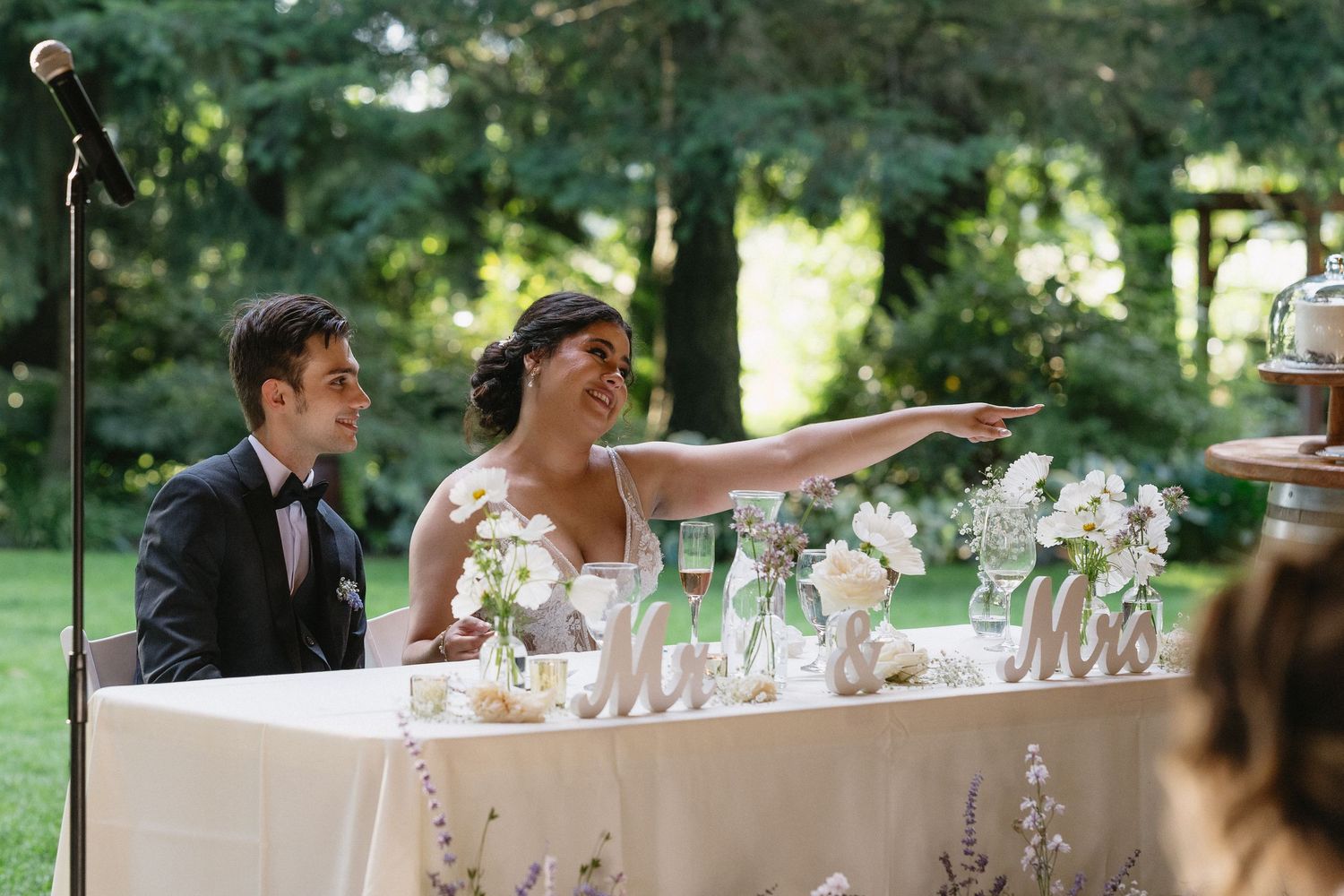 Smiling bride pointing while seated with groom at floral-decorated sweetheart table at outdoor wedding.