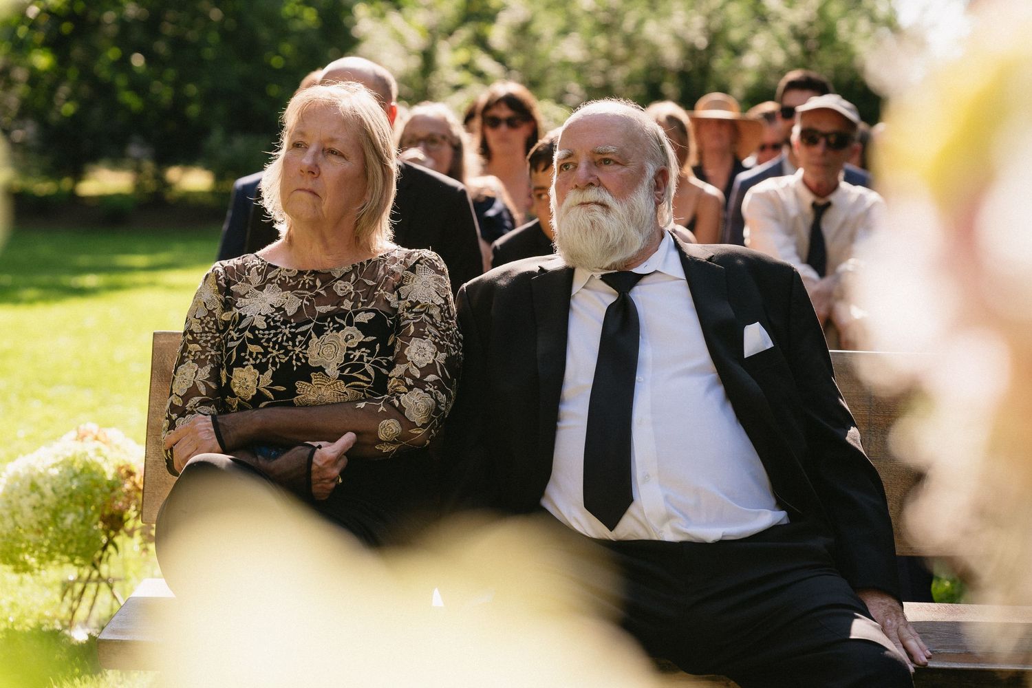 Elegant wedding guests seated outdoors watching ceremony, man in black suit with white beard and woman.