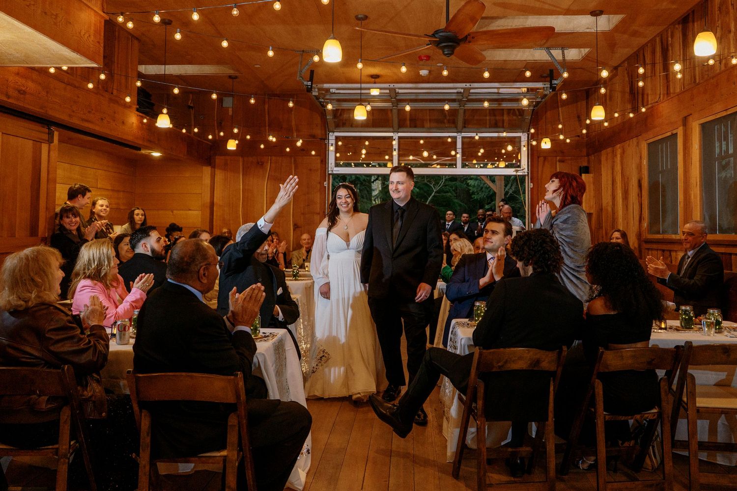 Newlywed couple making grand entrance into warmly lit rustic wedding reception hall with guests.