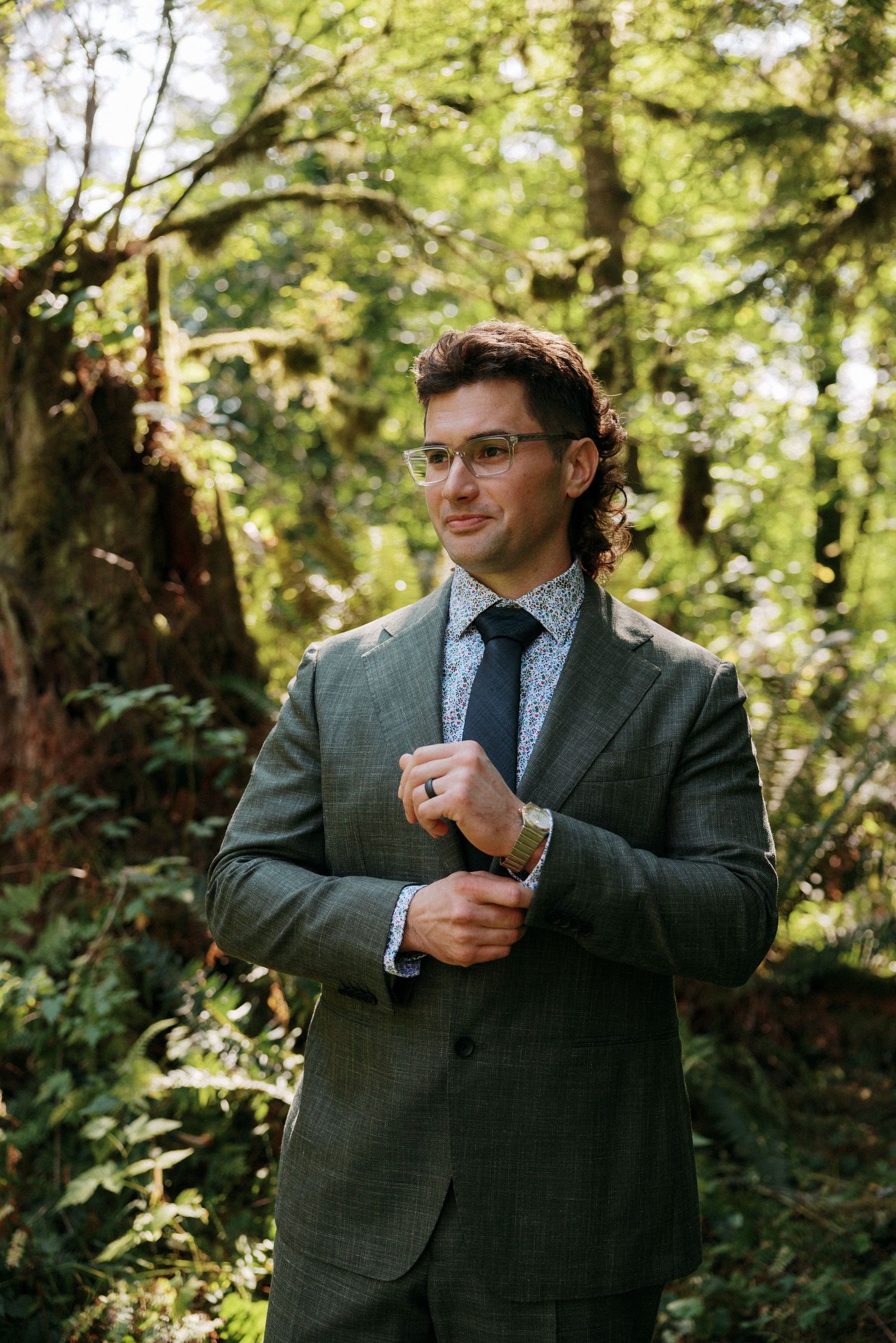 A well-dressed man in a gray suit and glasses adjusts his sleeve while standing in a lush forest.