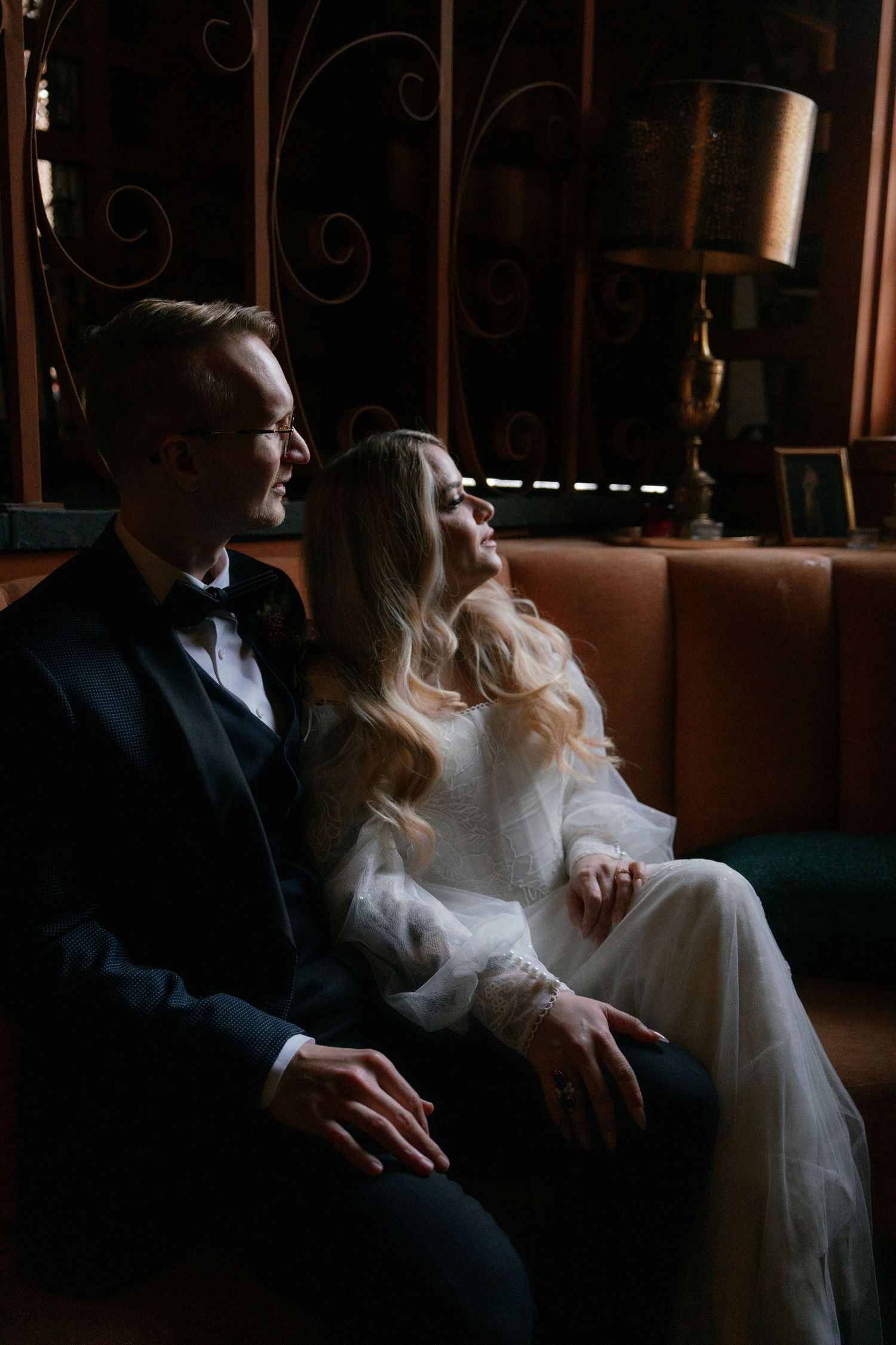 Bride and groom sitting together on vintage leather sofa in moody, warmly lit elegant wedding venue.