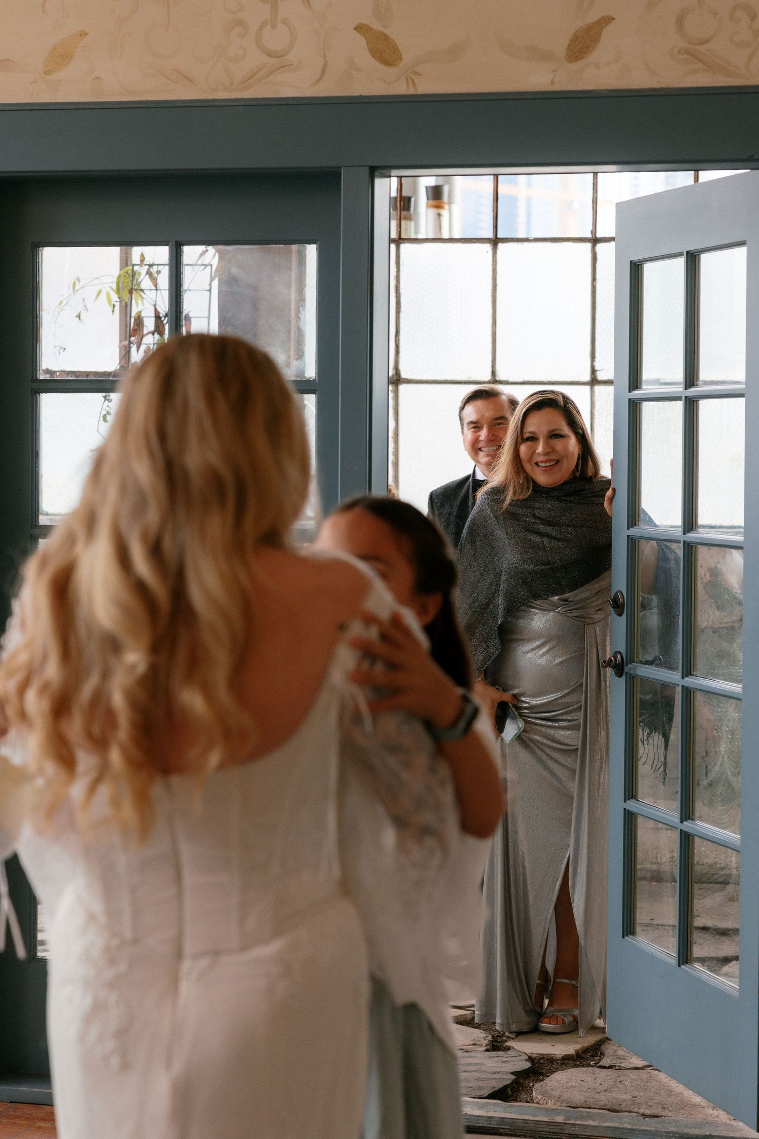 Bride hugs bridesmaid as smiling couple looks on through blue French doors at wedding.