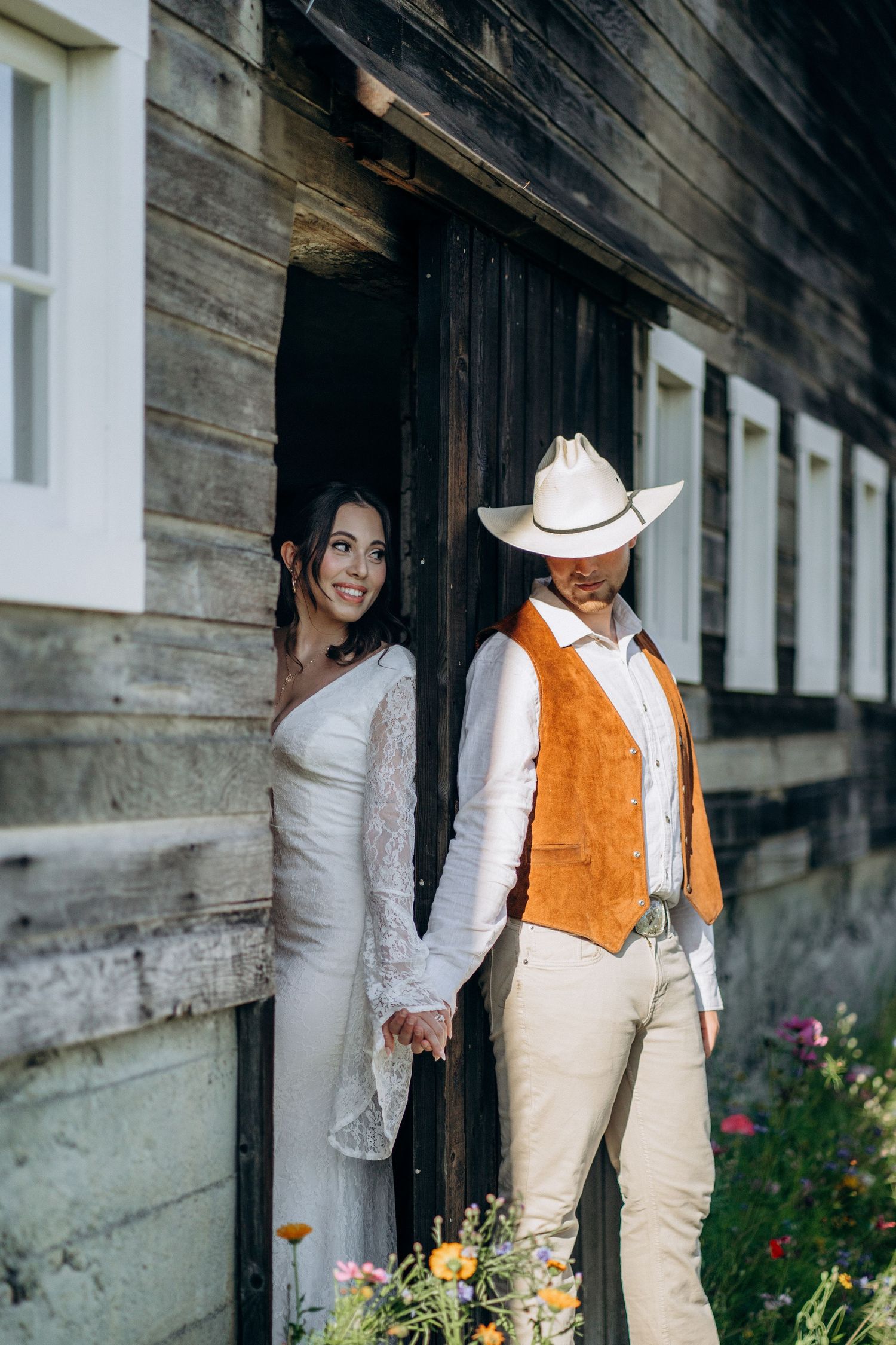 Smiling bride and cowboy-hat groom holding hands outside rustic wooden barn with wildflowers.