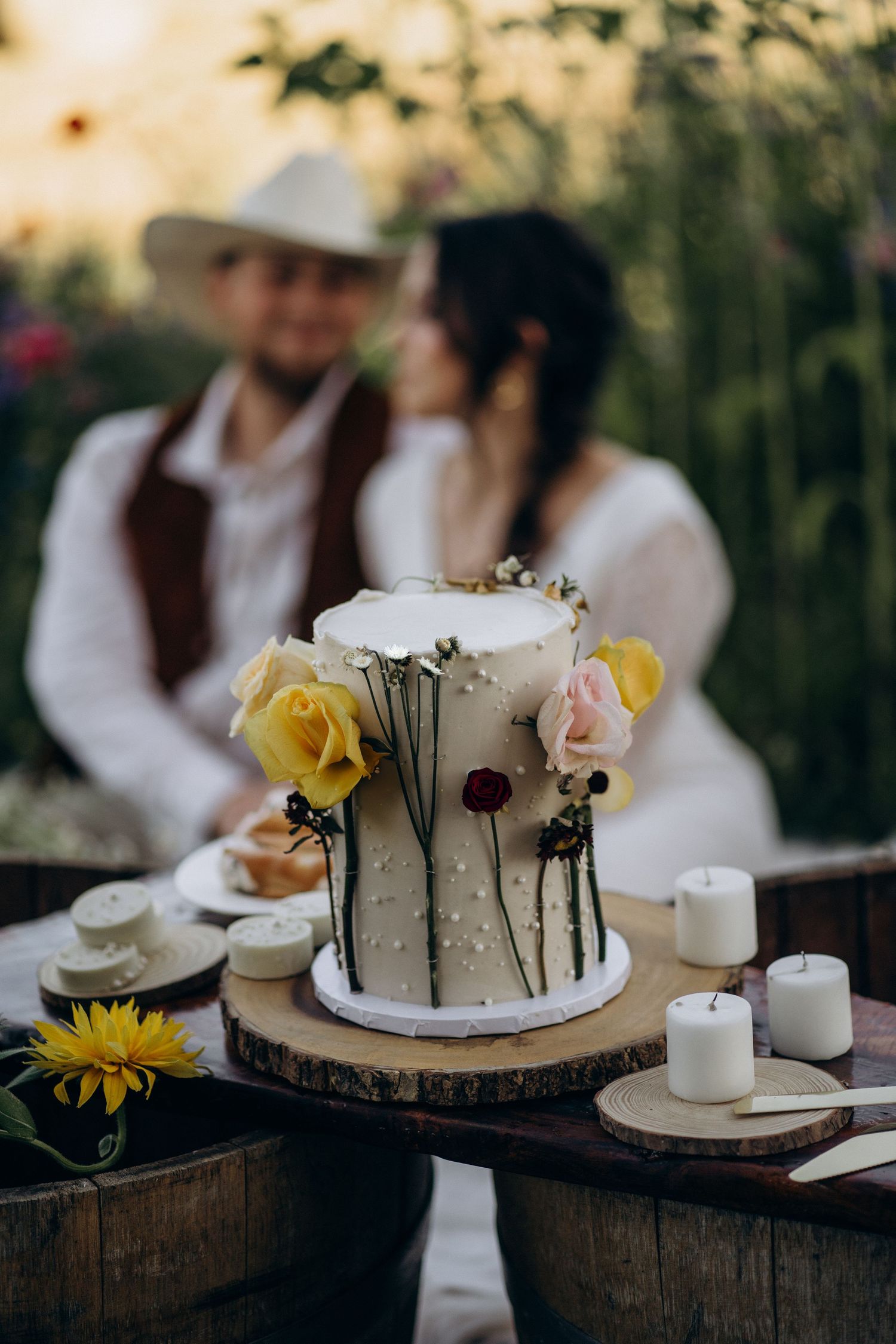 Elegant two-tier white wedding cake with roses on wood slice, couple embracing in flower field behind.